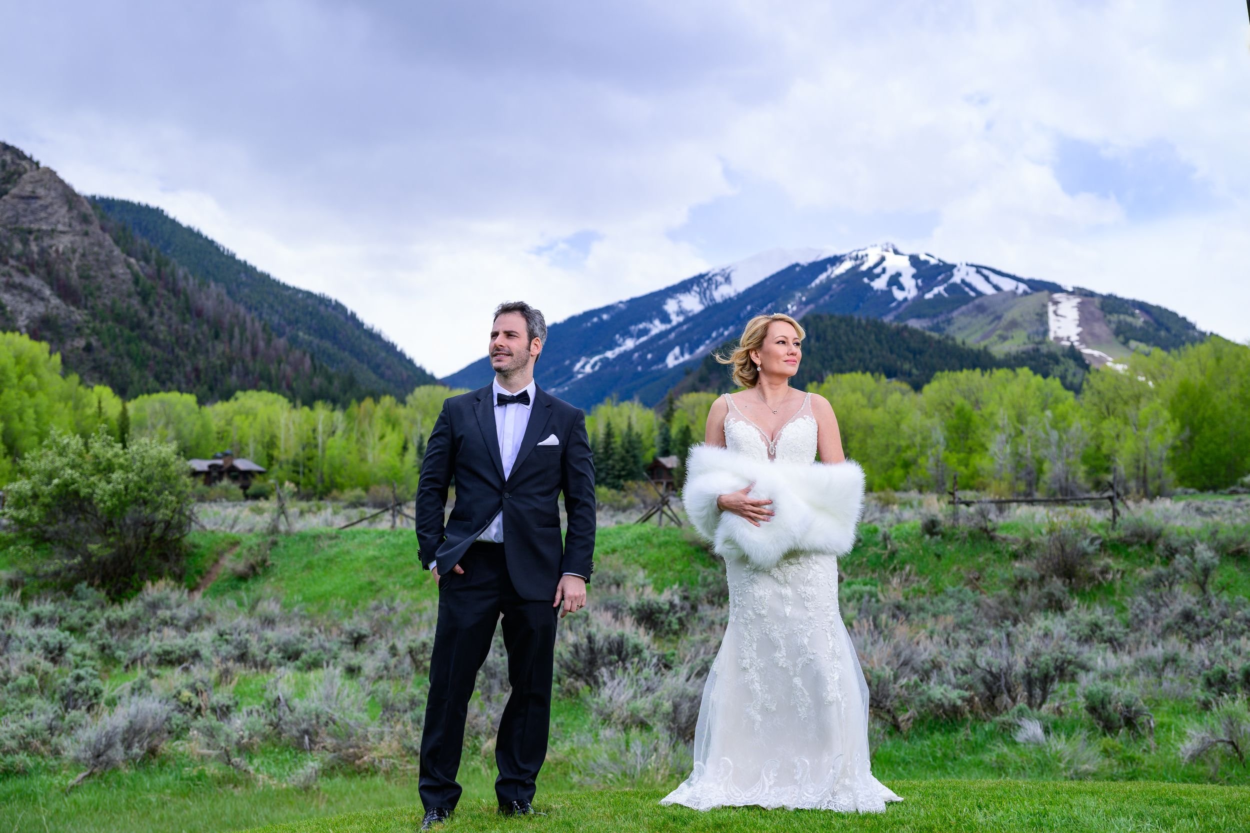 Bride and groom posing outdoors in formal attire with a mountain and greenery backdrop.