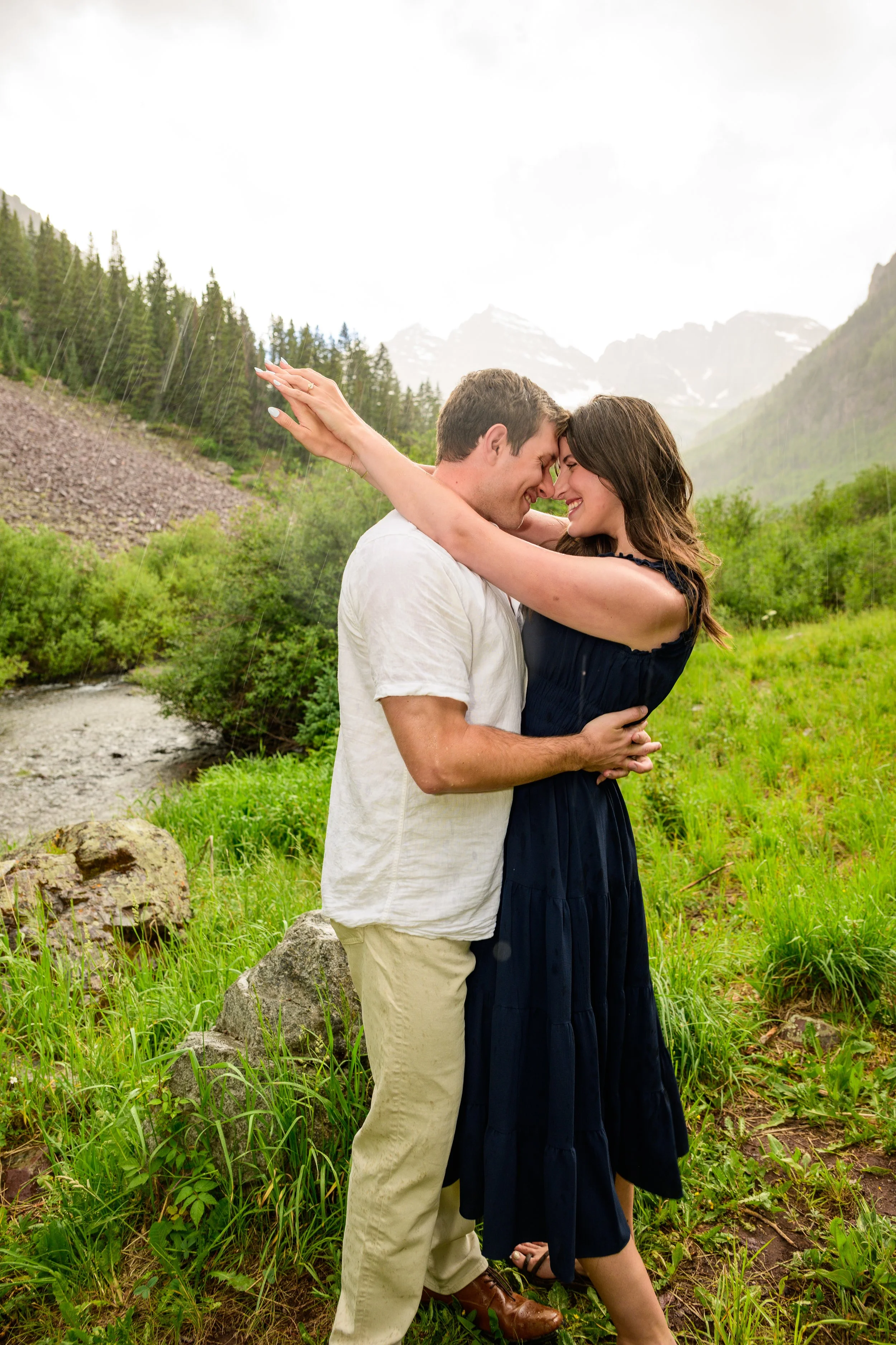 A couple stands close together, their foreheads touching, with a mountain landscape in the background during rain. The man wears a white shirt and beige pants, and the woman wears a dark blue dress. They appear happy and intimate, embracing each othe