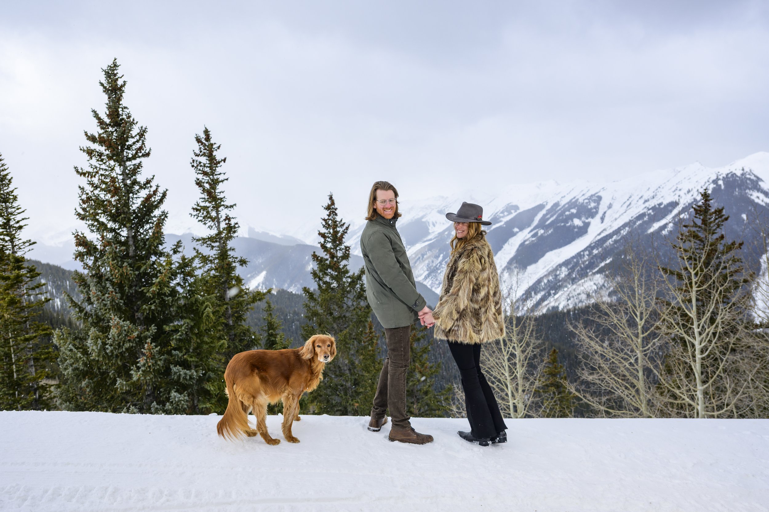 A couple holding hands in a snowy mountain landscape with their dog, snow-covered trees, and distant mountains in the background.