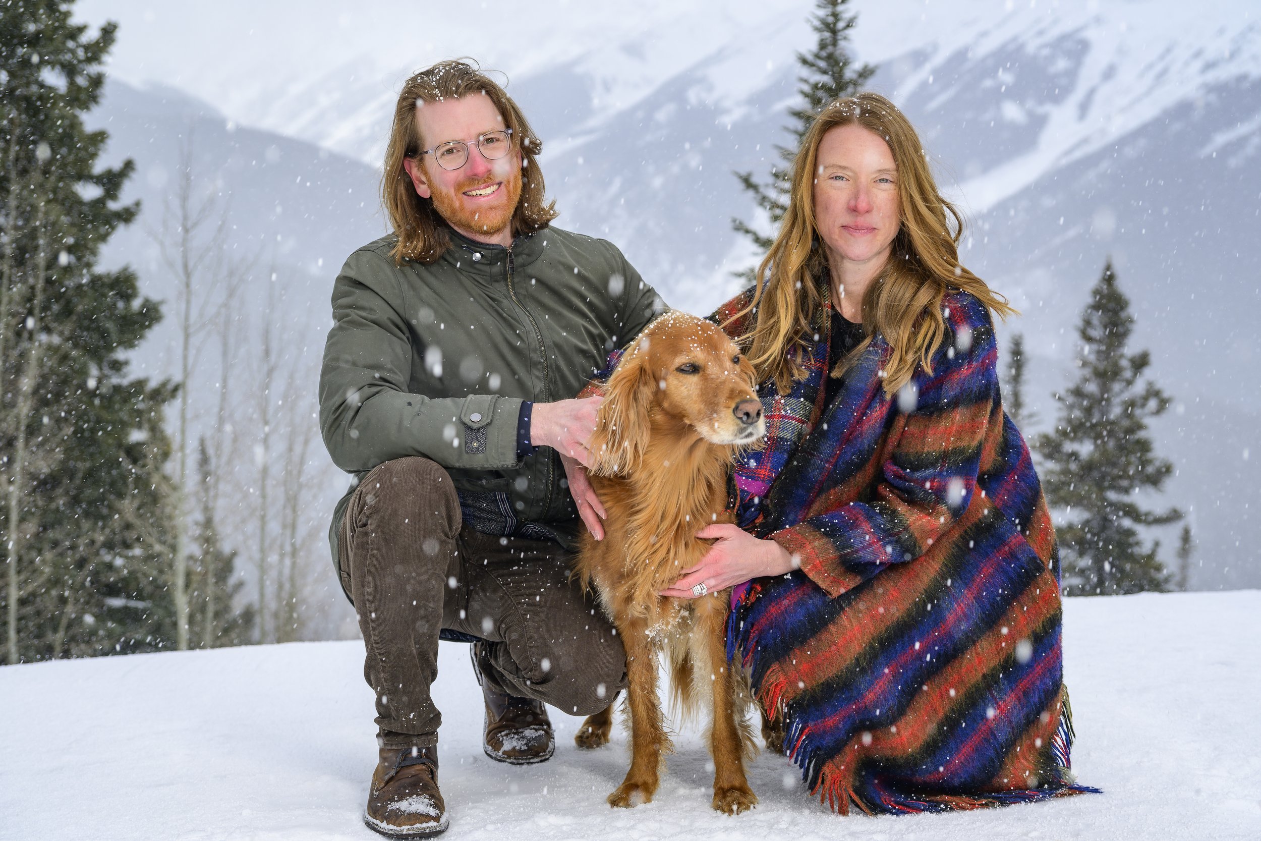 A couple and their golden retriever dog standing in a snowy outdoor landscape with snow-covered mountains and tall evergreen trees in the background.