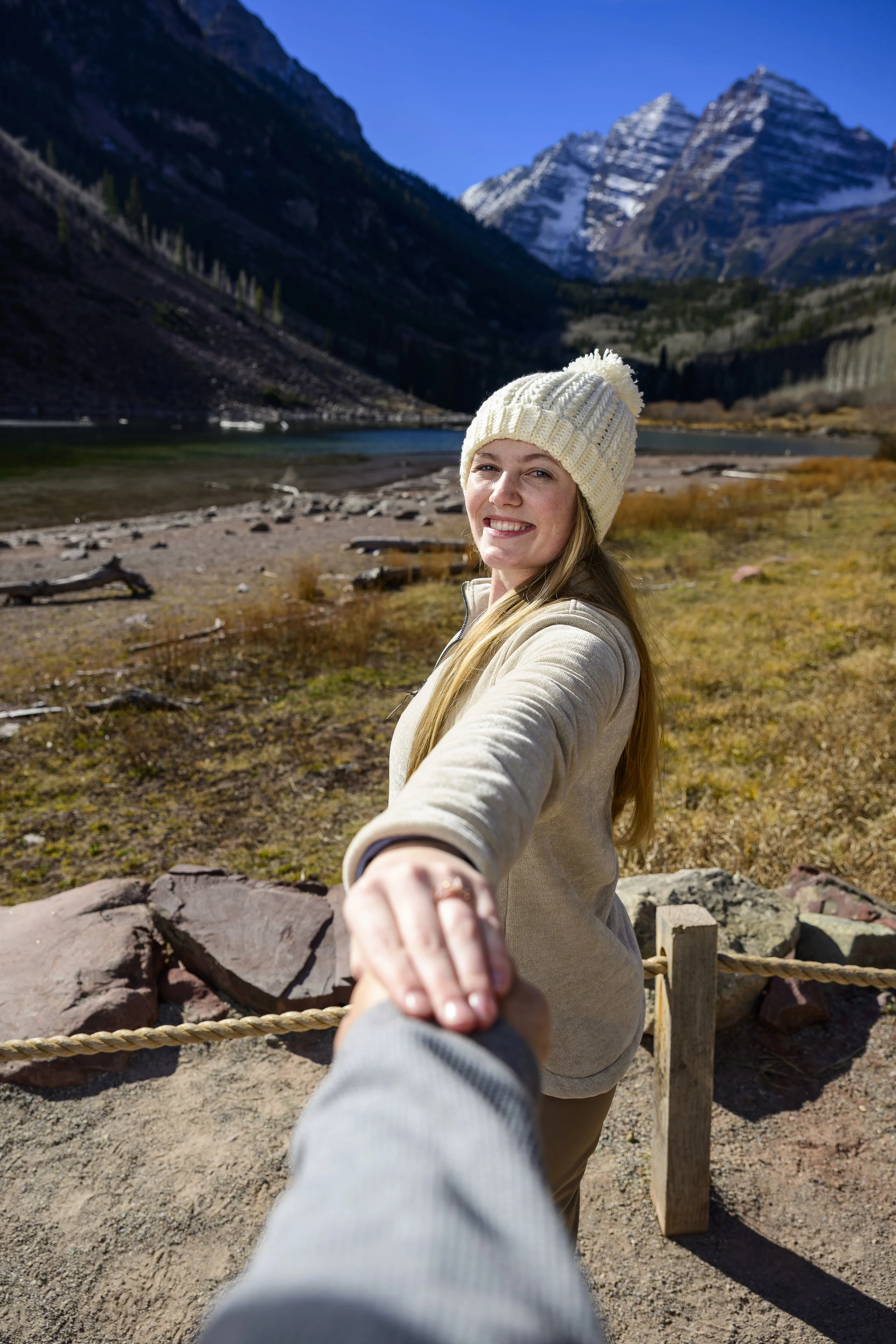A woman in a cream knit hat and beige jacket holding hands with the camera owner, standing outdoors near a mountain lake with snow-capped peaks in the background.