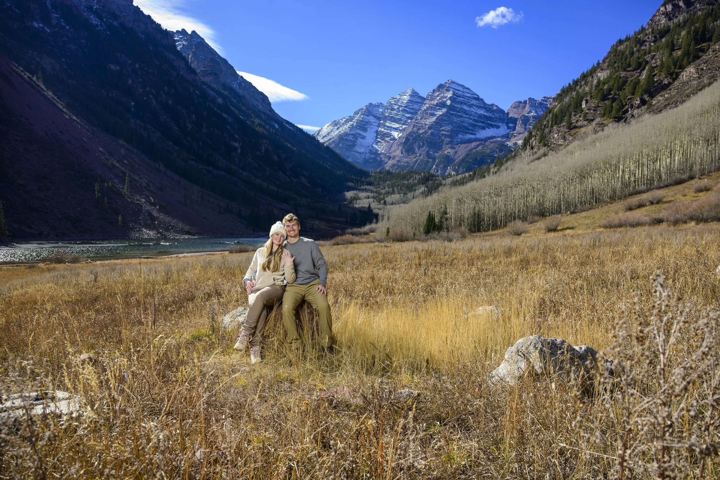 A couple sitting on a rock in a scenic mountain valley, surrounded by tall grass and mountains with snow-capped peaks in the background, under a blue sky.