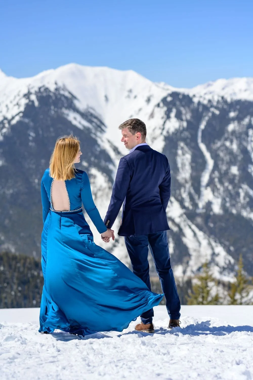 A couple standing on snow in front of a mountain landscape, dressed in formal attire; the woman in a blue gown and the man in a dark suit.