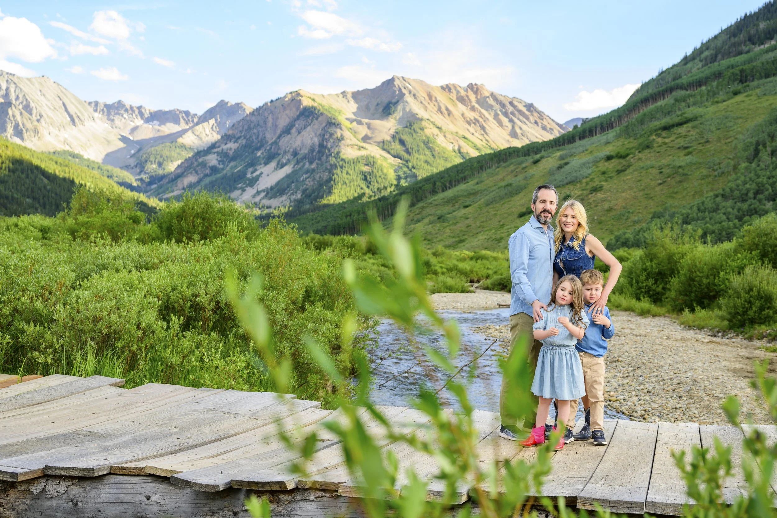 A family of four standing on a wooden bridge in a lush green valley with mountains in the background.
