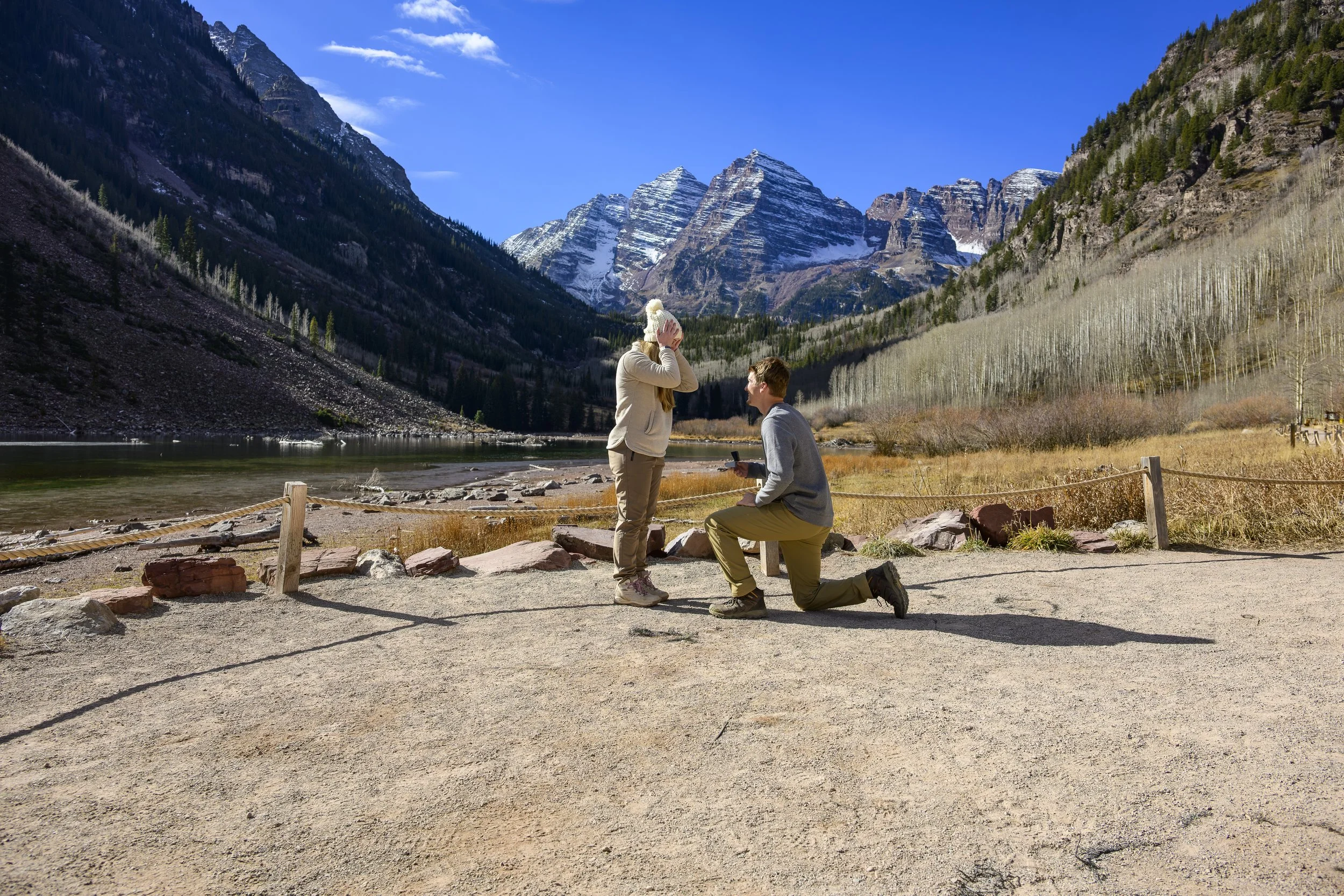A man proposing marriage to a woman by a lake with mountains in the background.