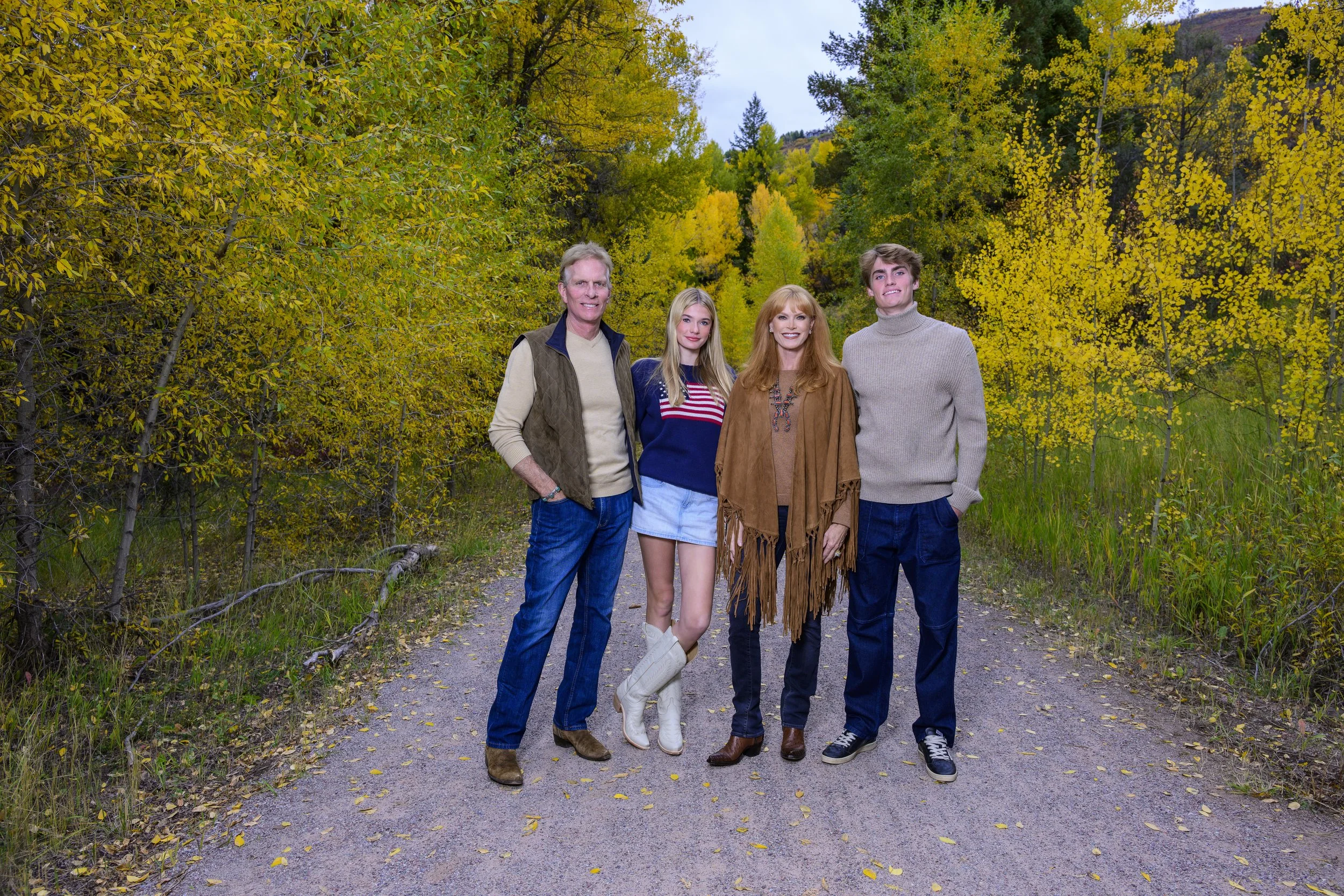 A family of five standing on a dirt path in a forest with fall foliage, including trees with yellow and green leaves. The family members are smiling and dressed in autumn attire.