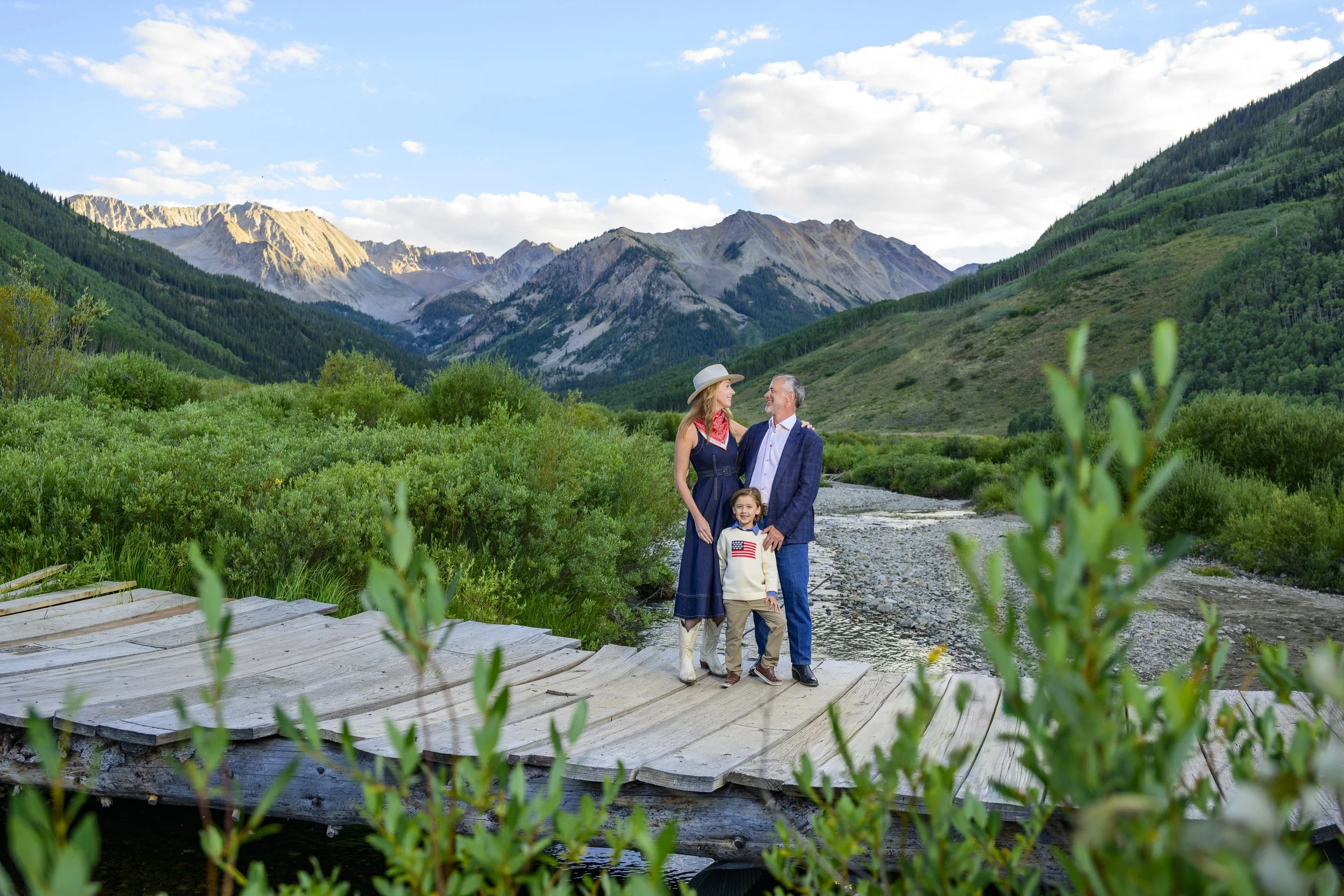 A family of three, a woman, a man, and a young girl, stands on a wooden bridge in a lush green landscape with mountains in the background, during daytime.