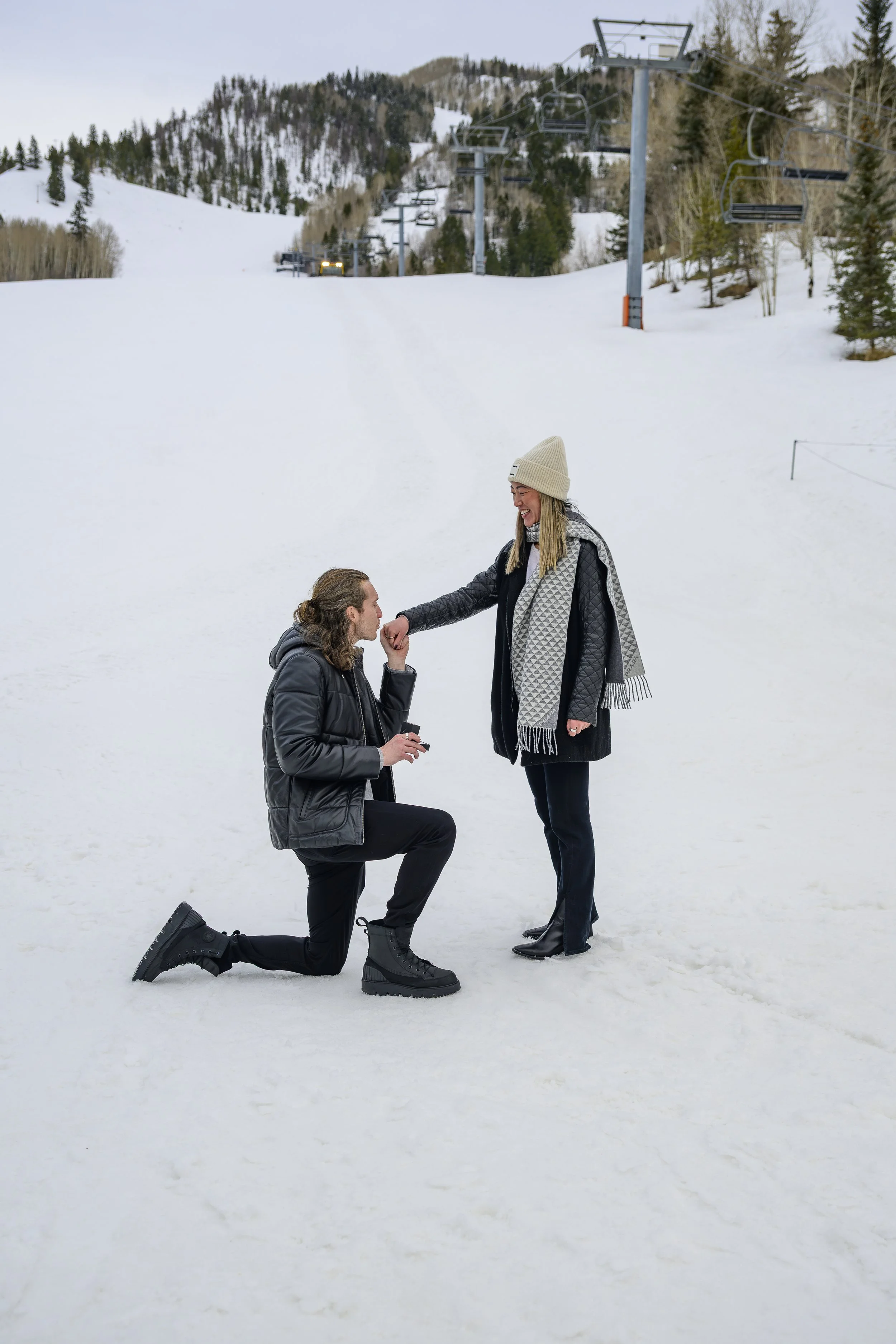 A man proposing to a woman in the snow at a ski resort, with ski lifts and snow-covered mountains in the background.