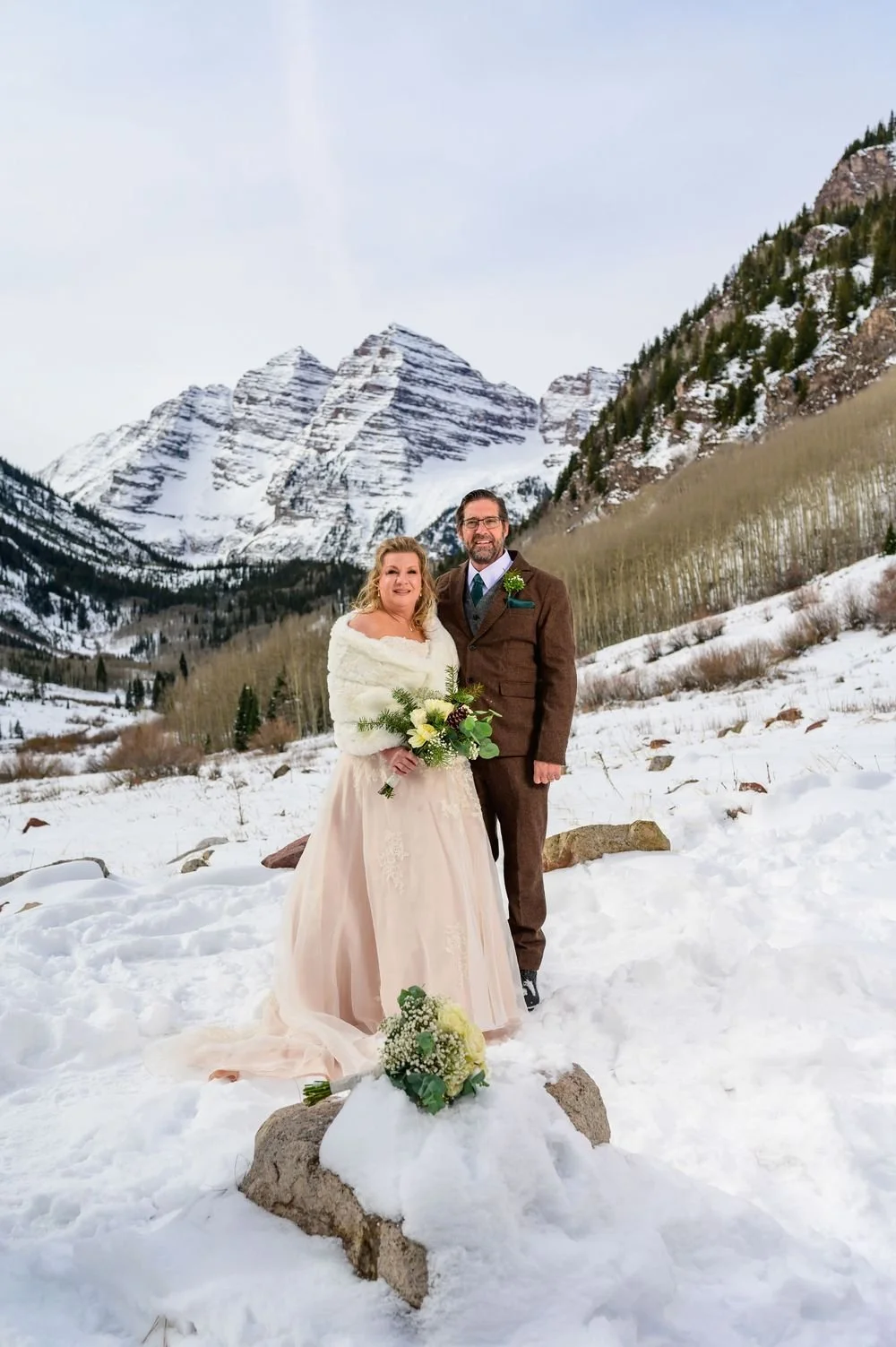 A couple in wedding attire posing in a snowy landscape with snow-covered mountains in the background.