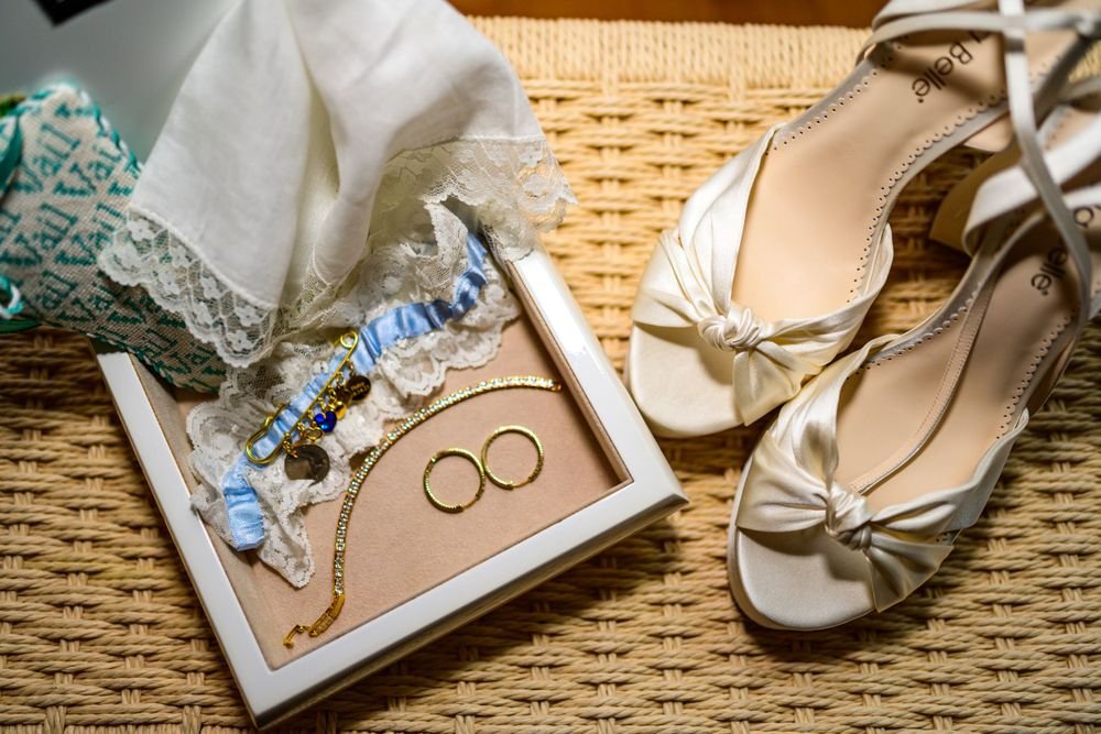 Pair of white bridal sandals next to a box containing lace garter, handkerchief, gold jewelry, and decorative items on a wicker surface.