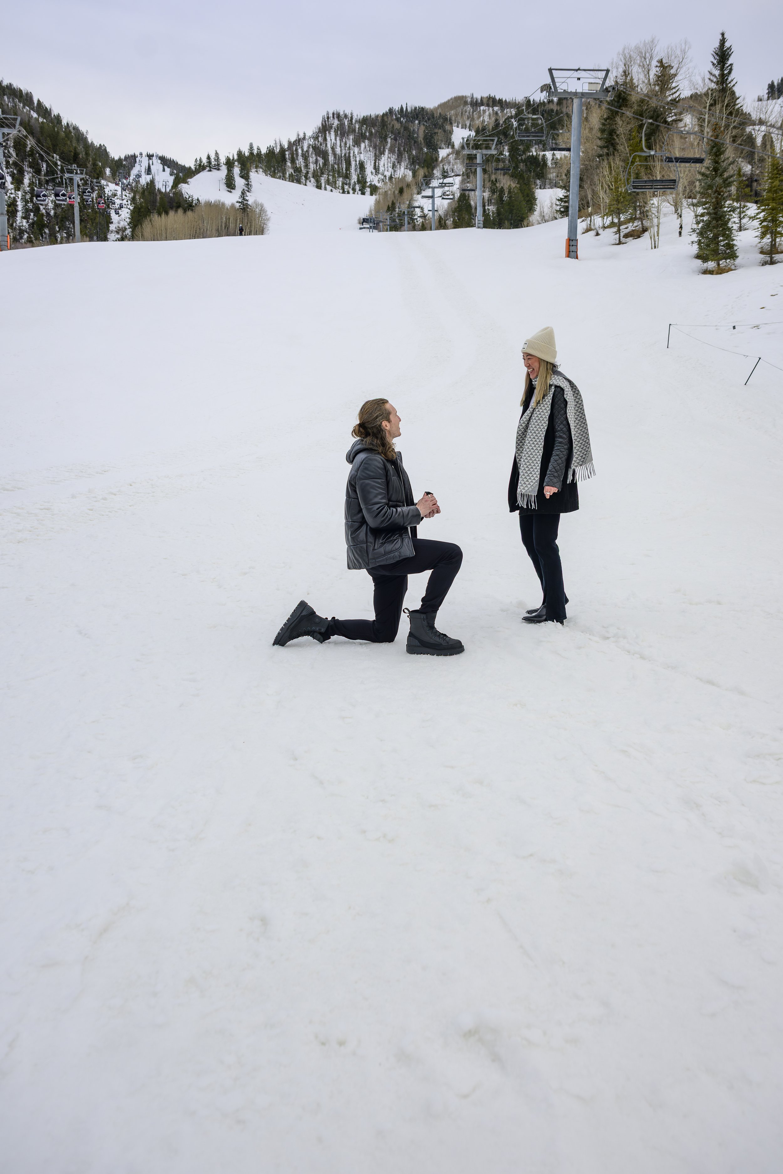 A man proposing marriage to a woman on a snowy ski slope with ski lifts and mountain scenery in the background.