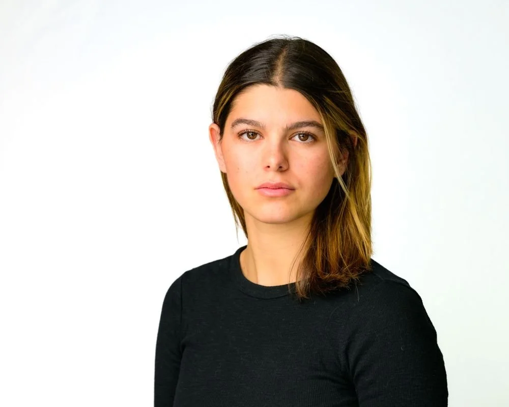 A young woman with long brown hair wearing a black top, looking directly at the camera against a plain white background.