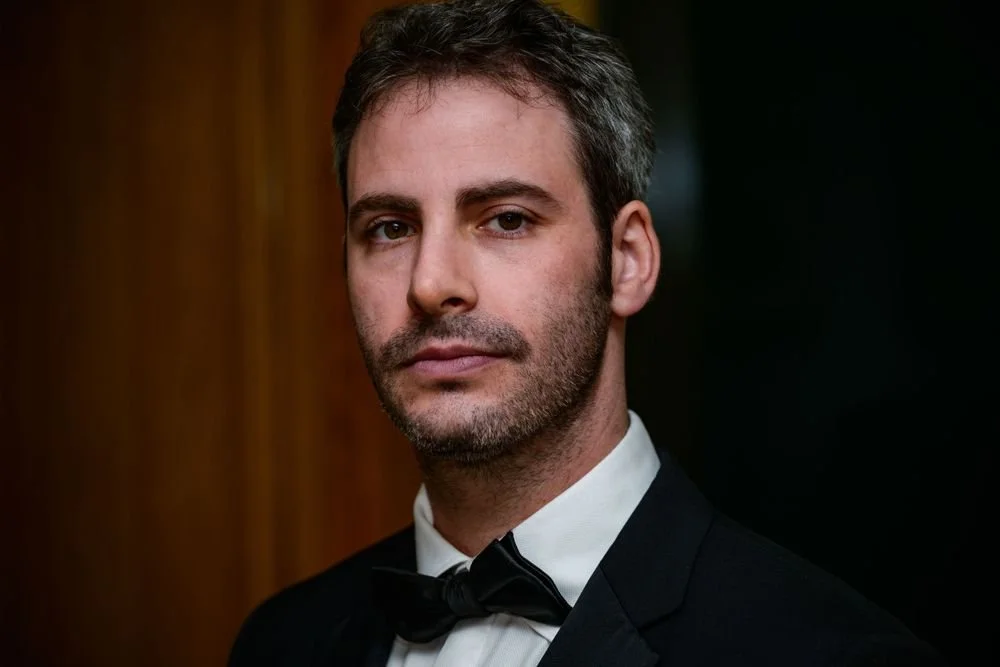 Man in formal attire with a bow tie, short hair, lightly bearded, looking at the camera; dark background.