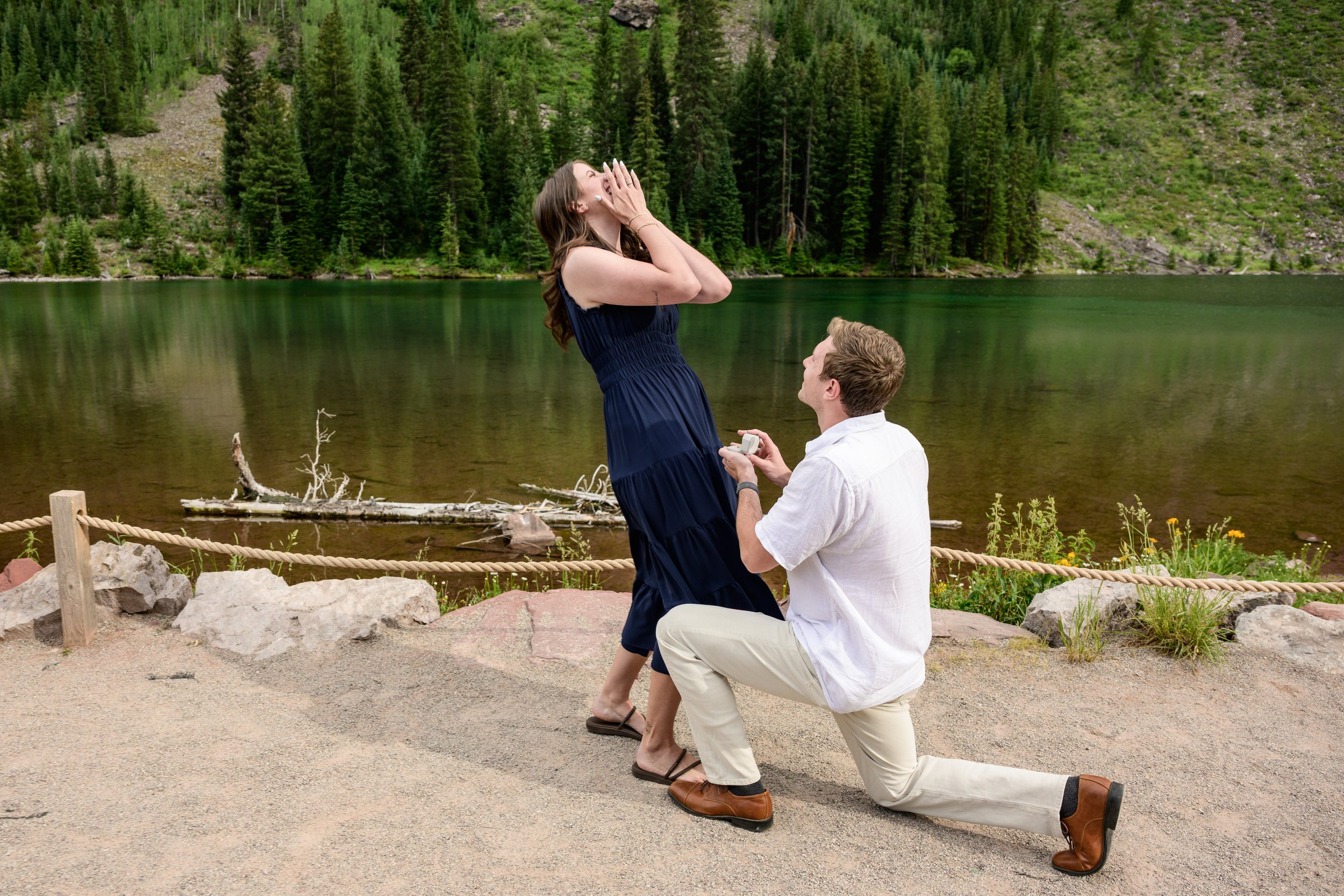 A man kneeling on one knee, holding a ring box and proposing to a woman near a lake surrounded by dense greenery.