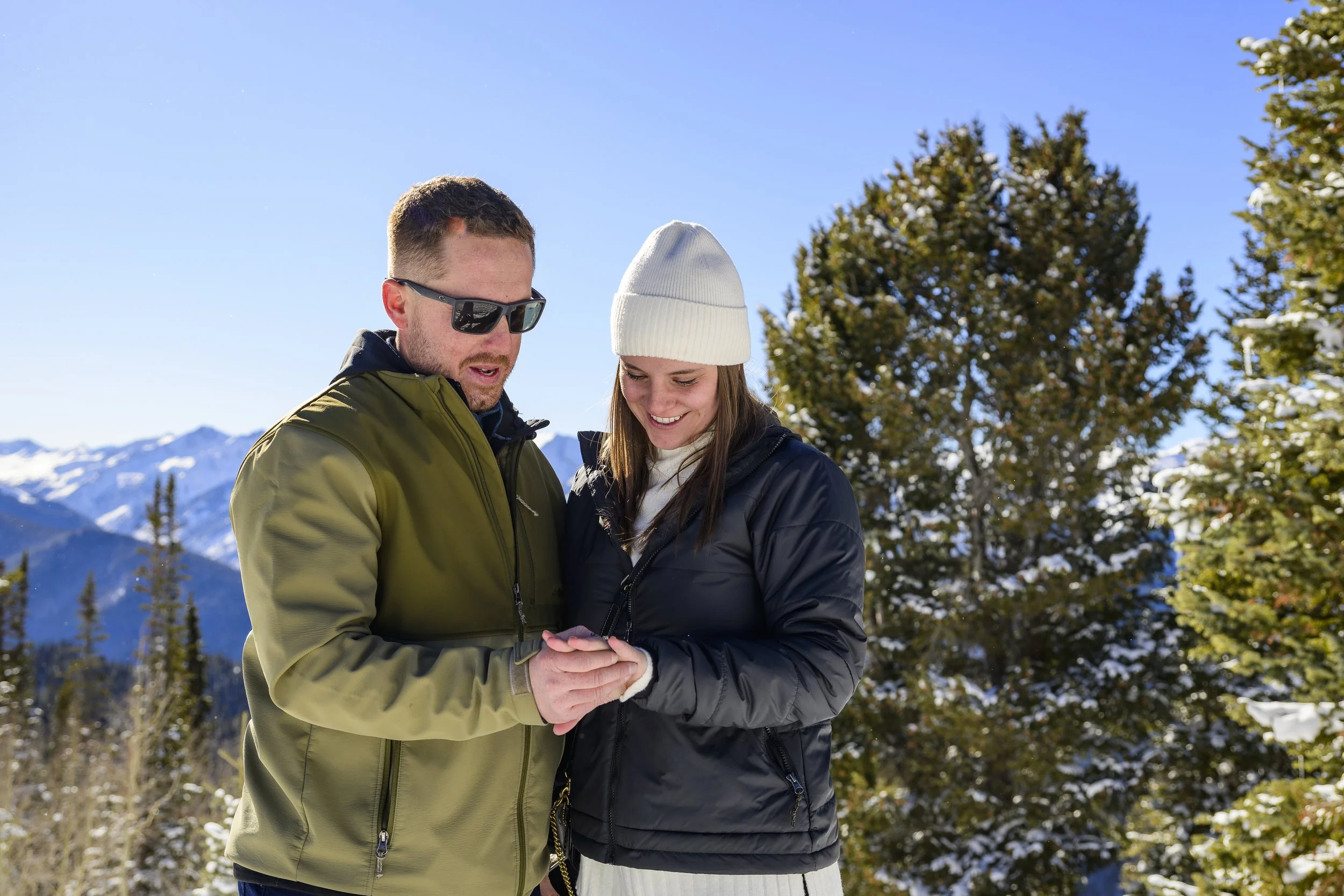 A man and woman stand together outdoors in a snowy mountain landscape, looking at a phone and smiling.
