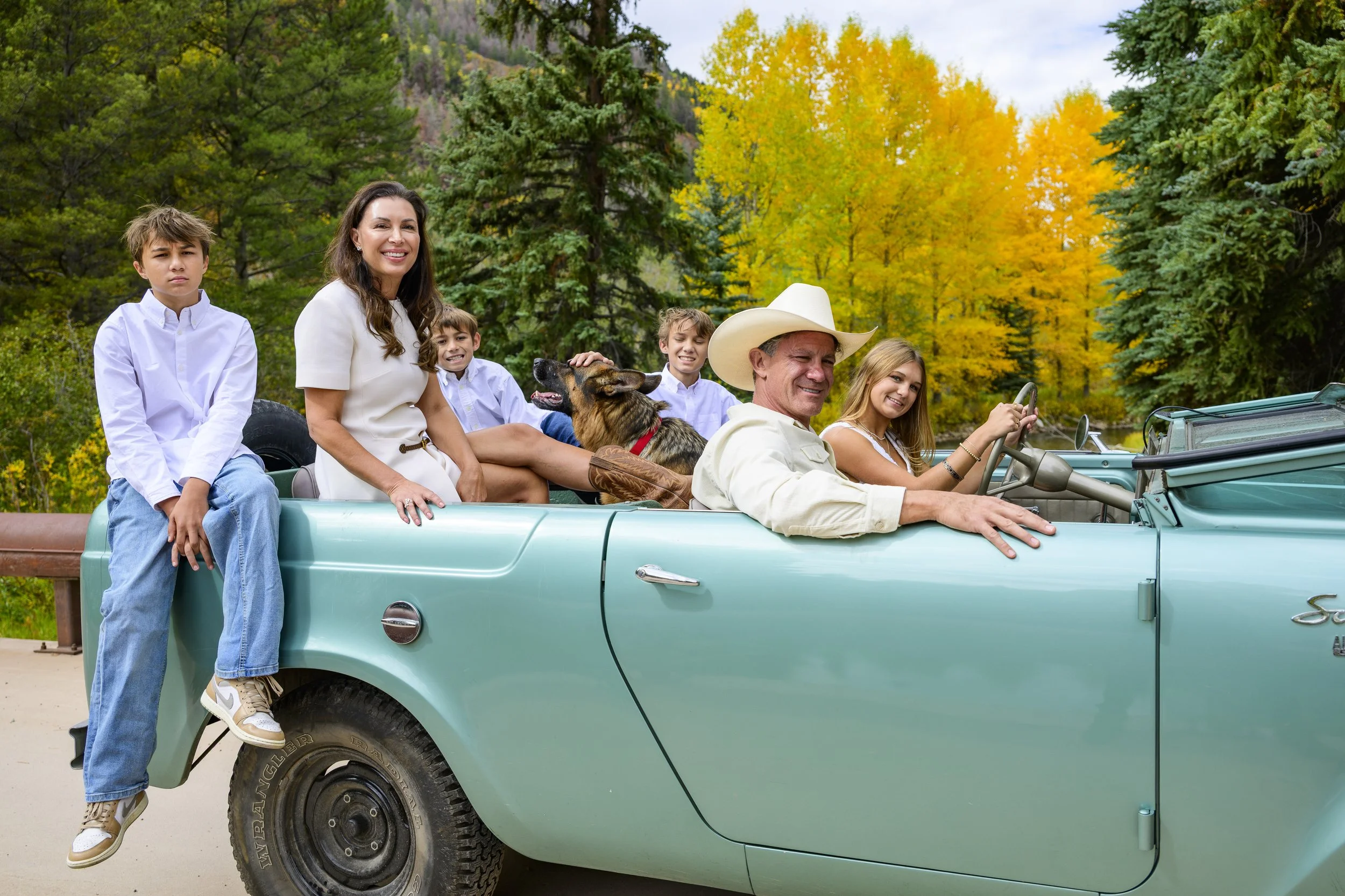 A family of six, including children, a woman, and a man wearing a cowboy hat, along with a dog, riding in a vintage turquoise convertible car on a scenic road surrounded by colorful autumn trees.