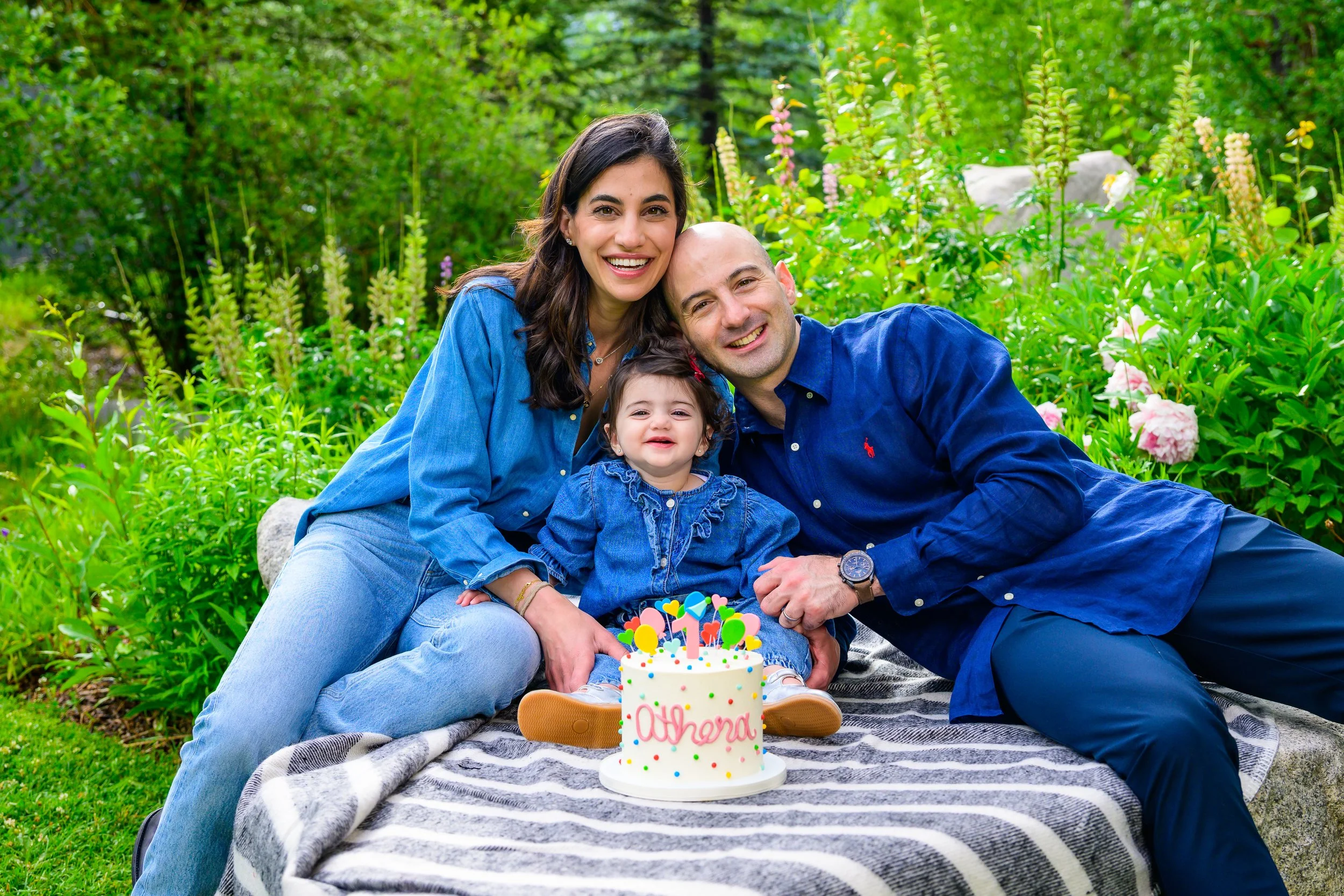 A family of three, a woman, a man, and a young girl, celebrating a birthday outdoors with a small cake that has birthday candles and colorful decorations.
