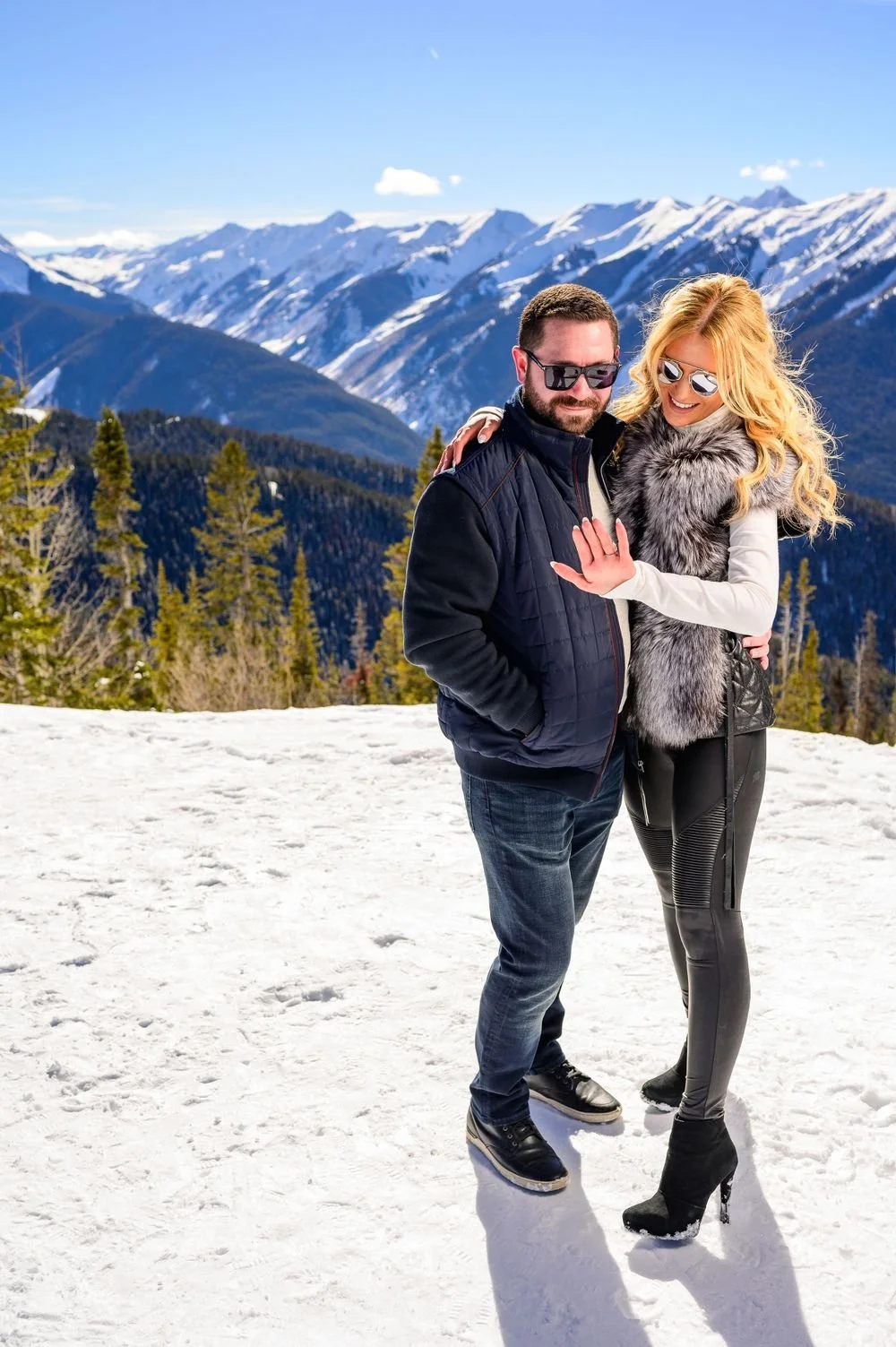 A couple in winter clothing standing on a snowy mountain with a scenic view of snow-capped peaks in the background.