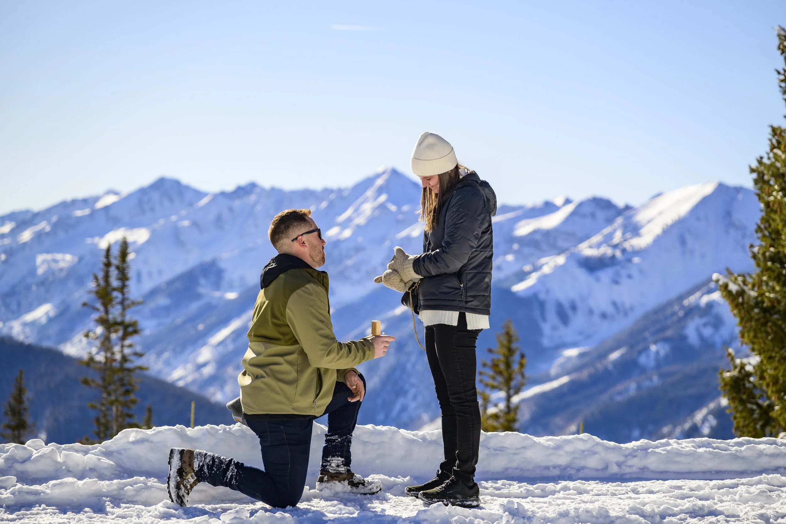 A man on one knee proposing to a woman in a snowy mountain landscape.