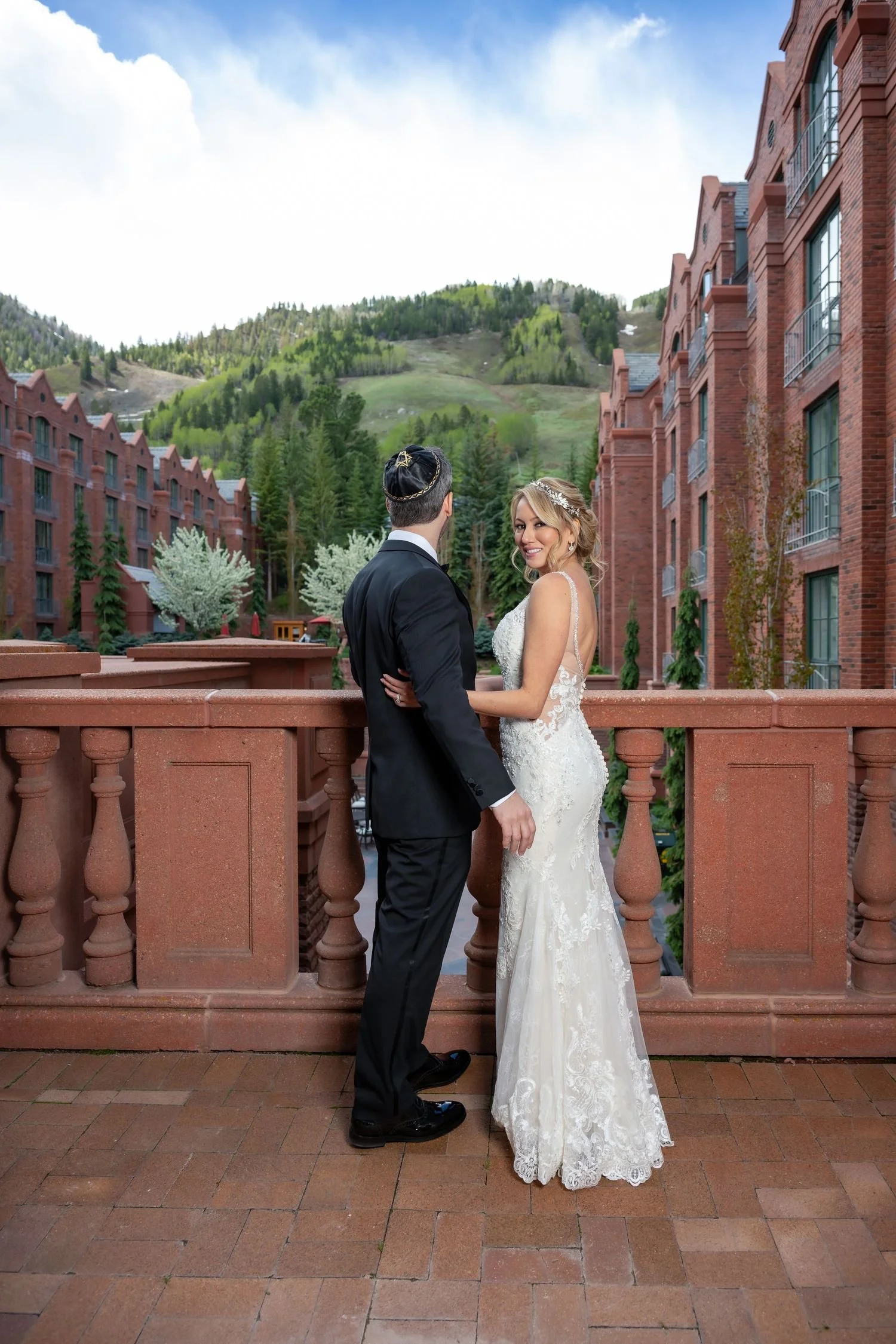 Bride and groom standing on a terrace with scenic mountain and forest view, red brick buildings in the background.