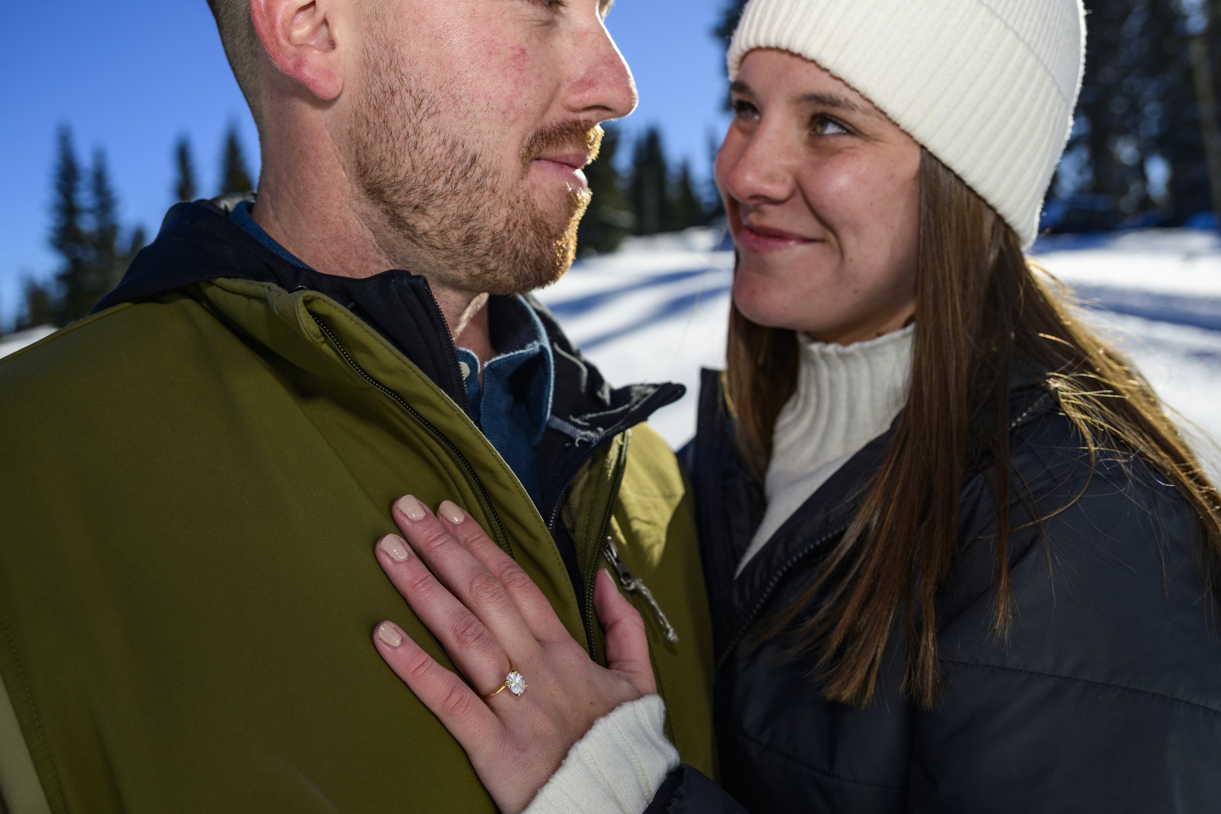 A woman with long brown hair wearing a white beanie and sweater, smiling at a man in a green jacket who is looking at her, outdoors in a snowy, forested area.