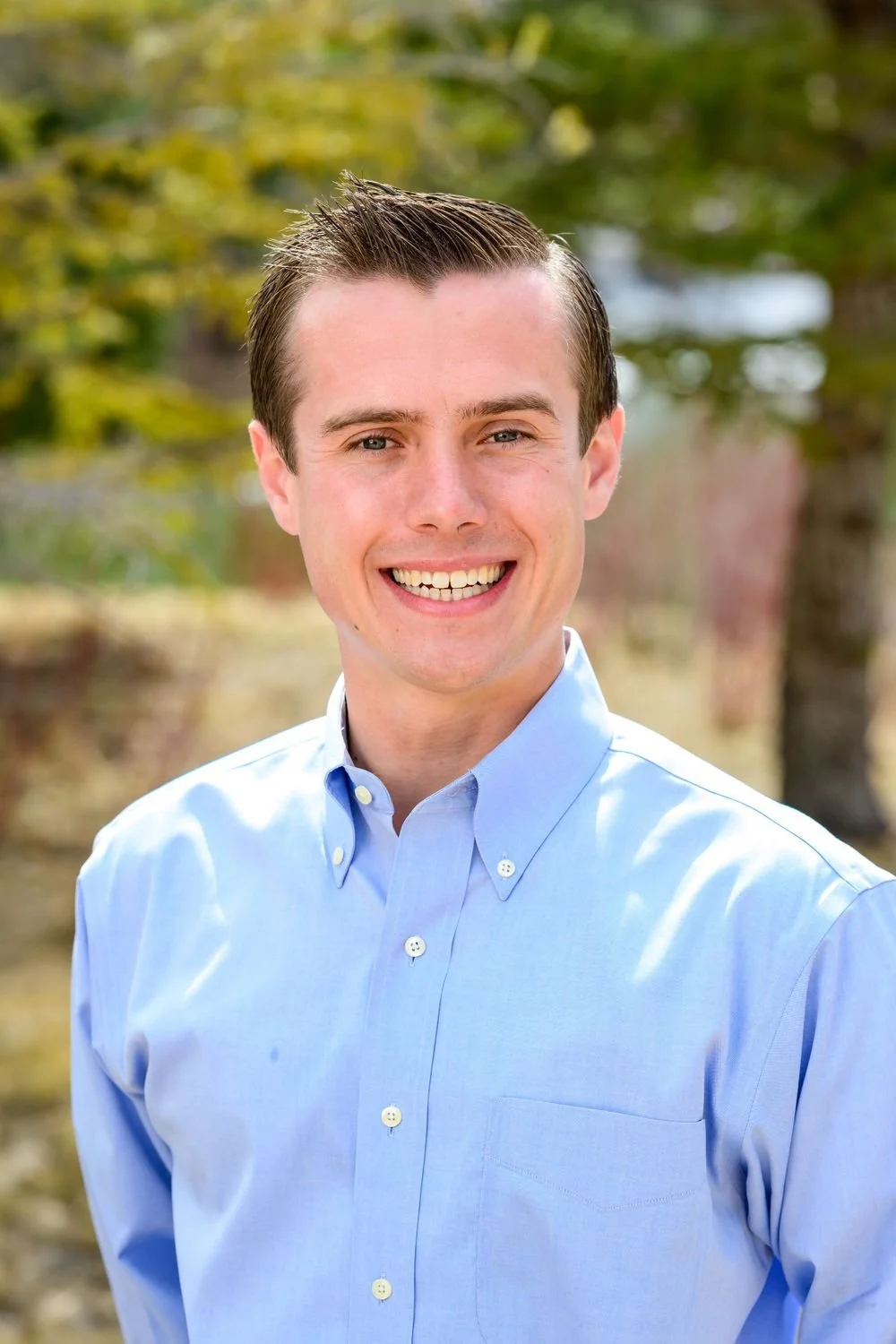 Man in a light blue button-up shirt smiling outdoors with greenery in the background.
