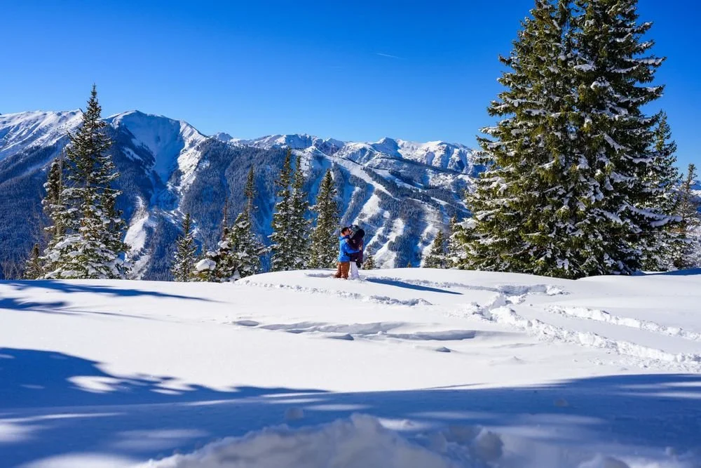 Snowy mountain landscape with a couple hugging, surrounded by pine trees and pristine white snow.