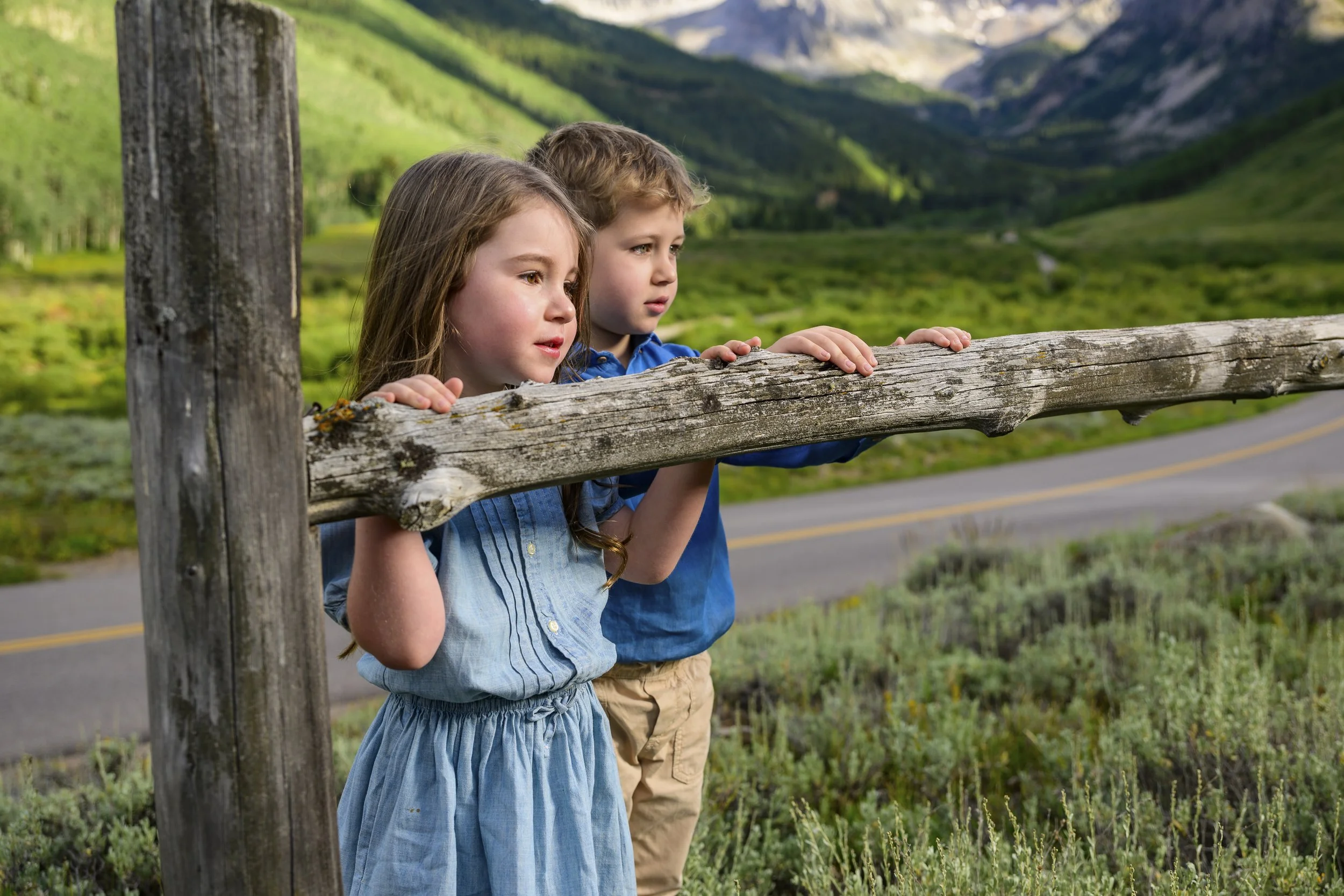 Two children, a girl and a boy, stand behind a wooden fence on a rural road, looking into the distance with a mountainous landscape in the background.