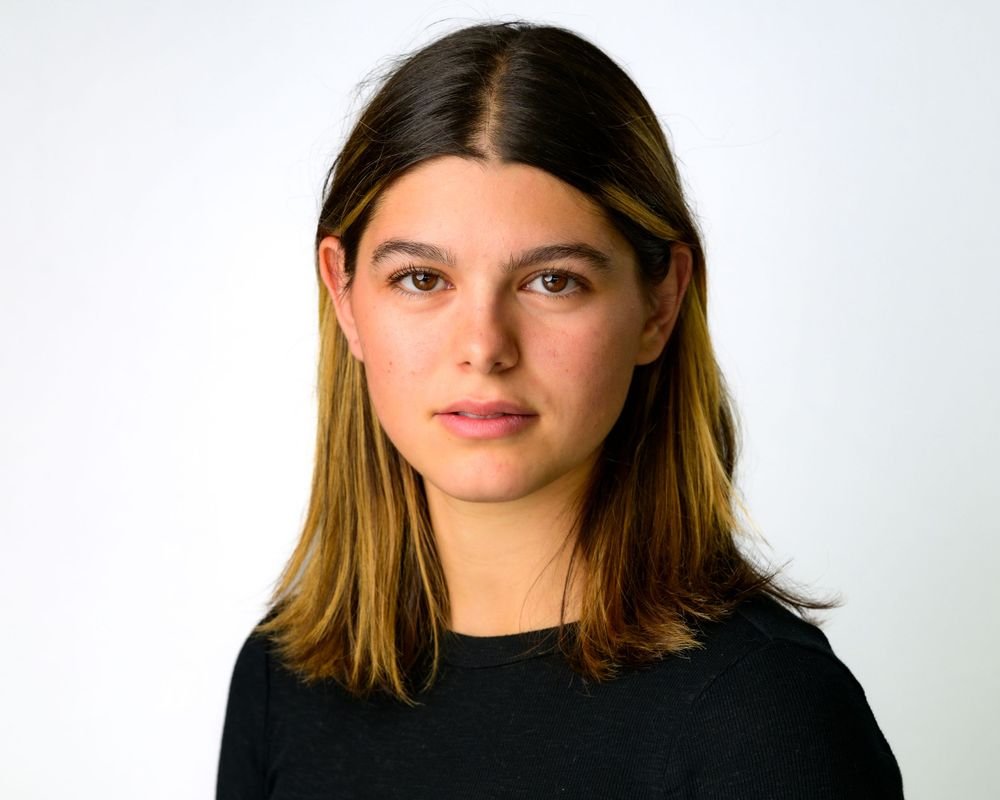 Young woman with long brown hair wearing a black shirt, looking directly at the camera, against a plain white background.