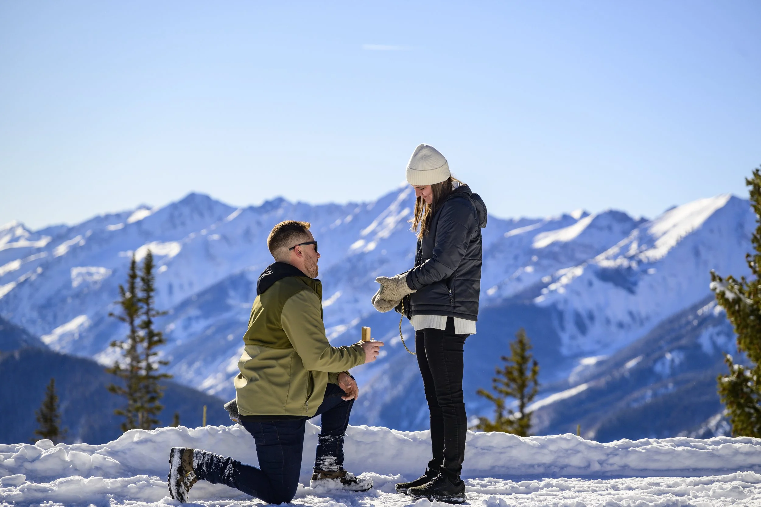 Man proposing to woman in snowy mountain setting with mountains in background.