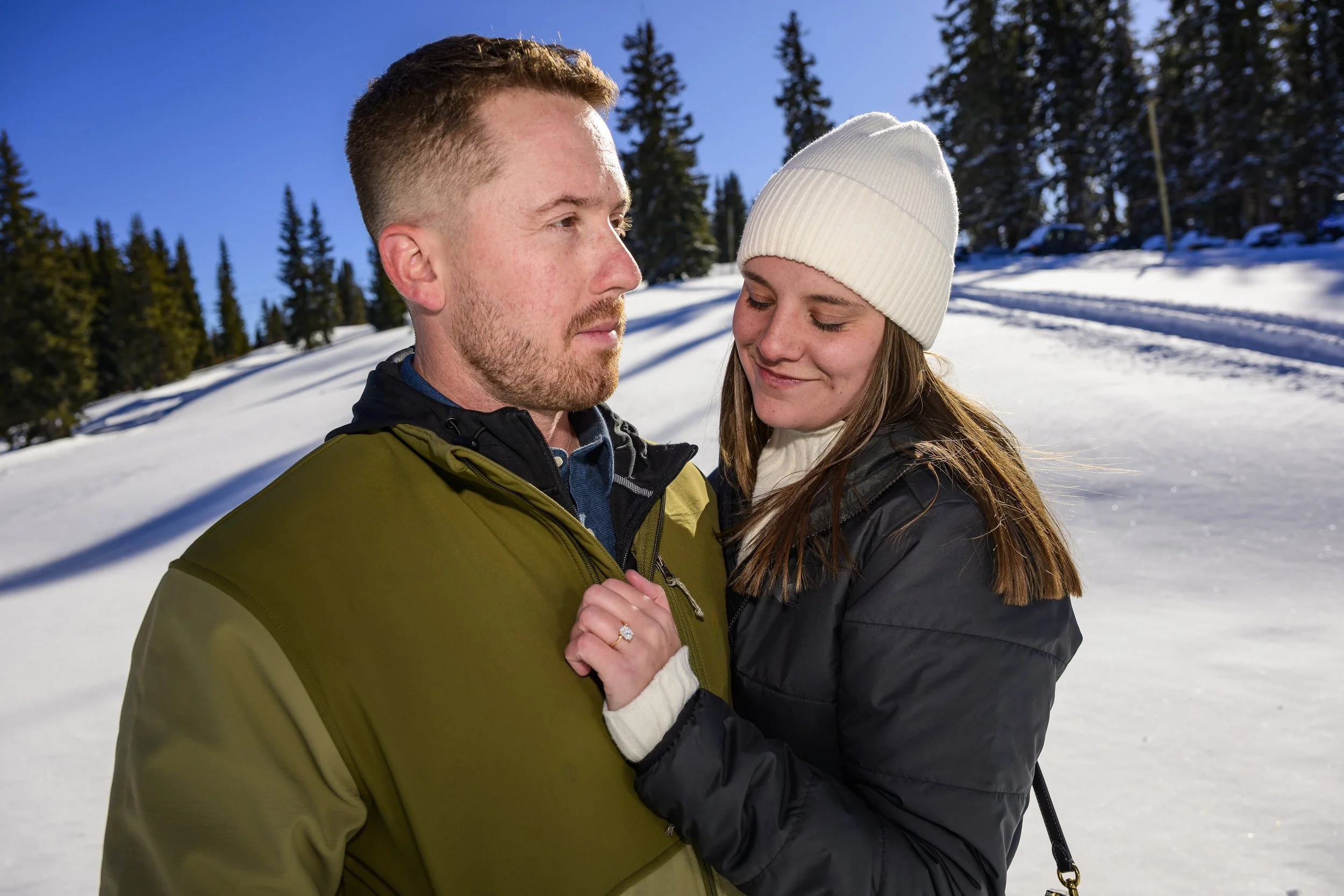 A young couple outdoors in winter, close together on snow-covered ground with trees in the background. The woman is smiling with her eyes closed, wearing a white beanie and black jacket. The man is looking down at her, wearing a green and black winte