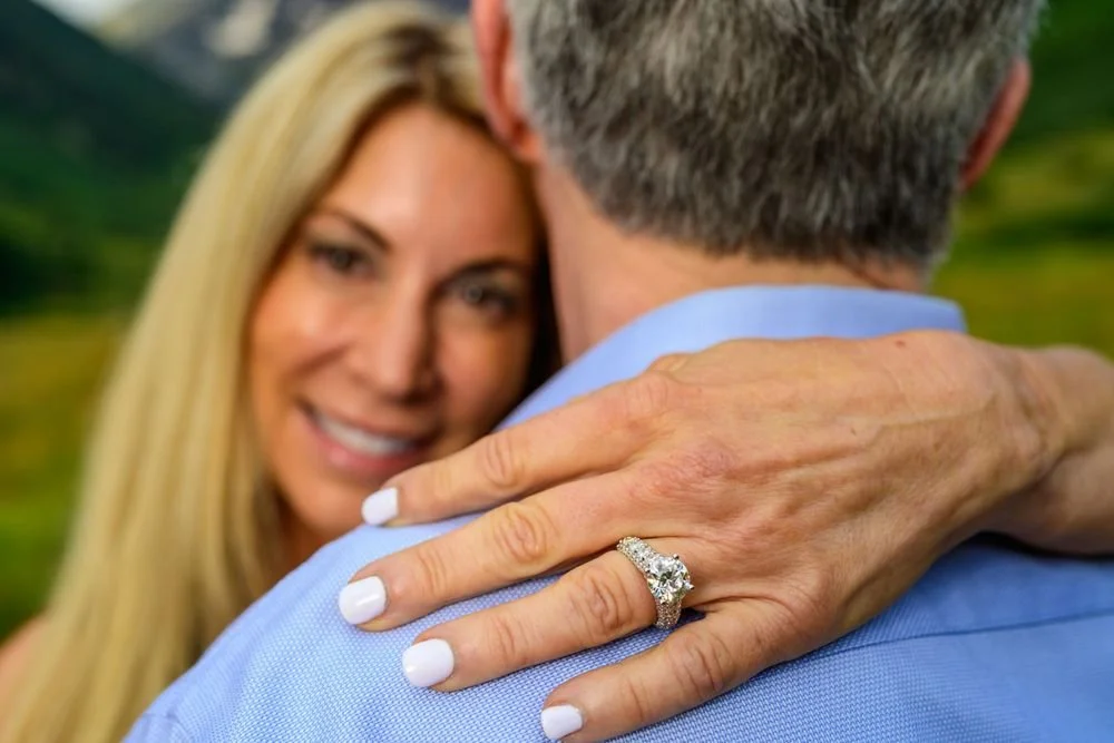 Close-up of a woman's hand with a large diamond ring on a man's shoulder, with the woman smiling in the blurred background.