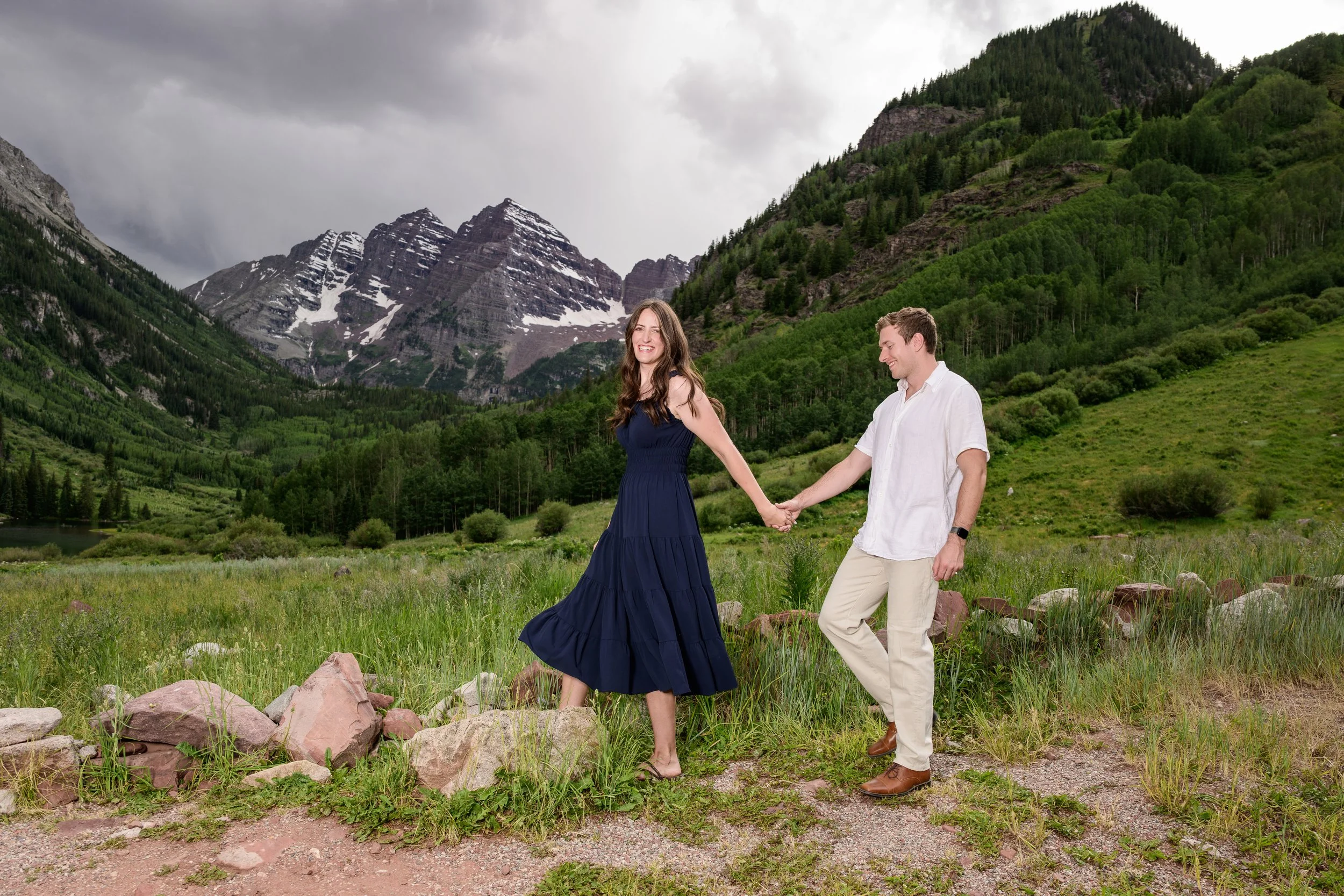 A couple holding hands and walking on a trail in a lush green mountainous area with snow-capped peaks in the background, under a cloudy sky.
