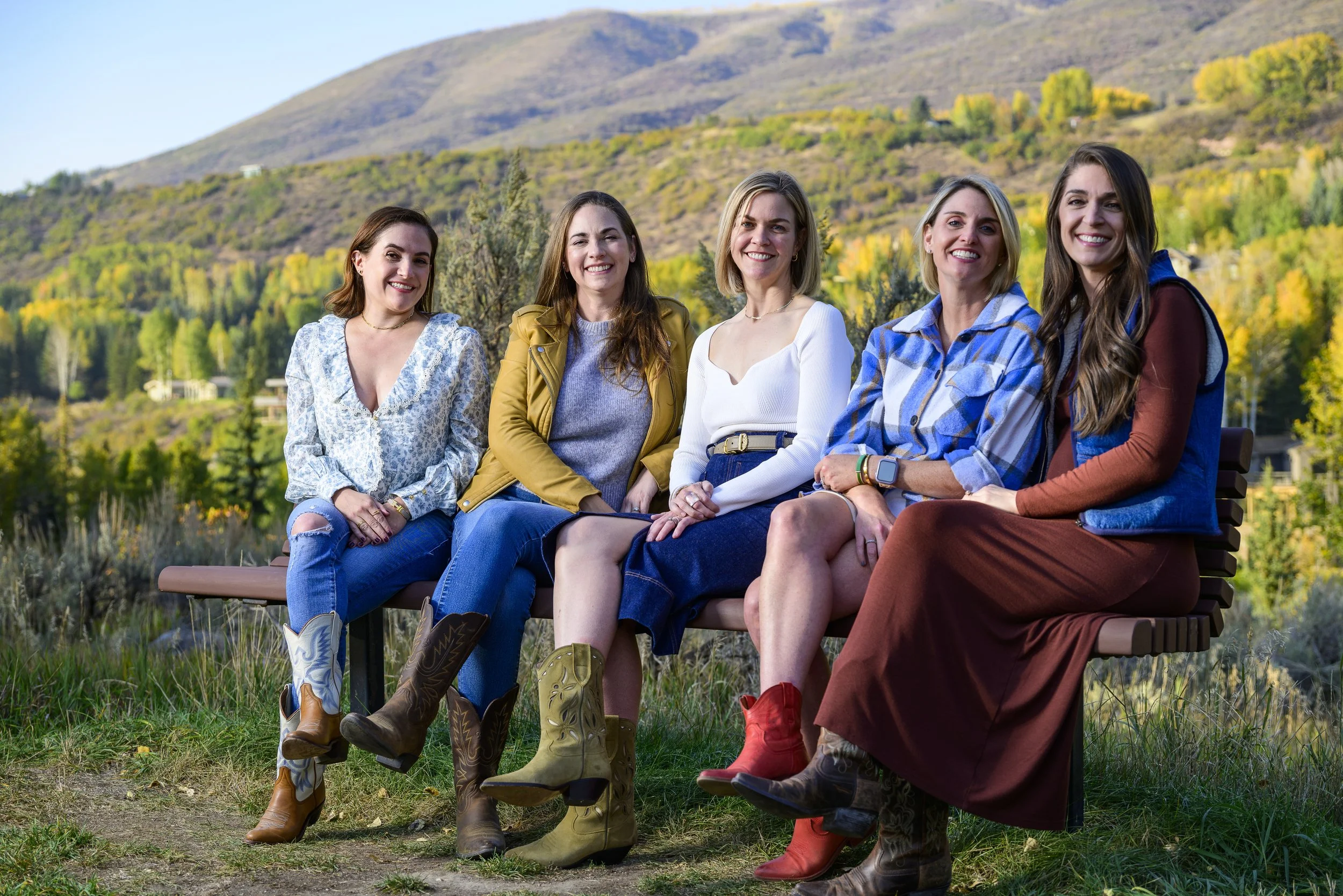 Five women sitting on a bench outdoors with a mountain and trees in the background, during autumn.