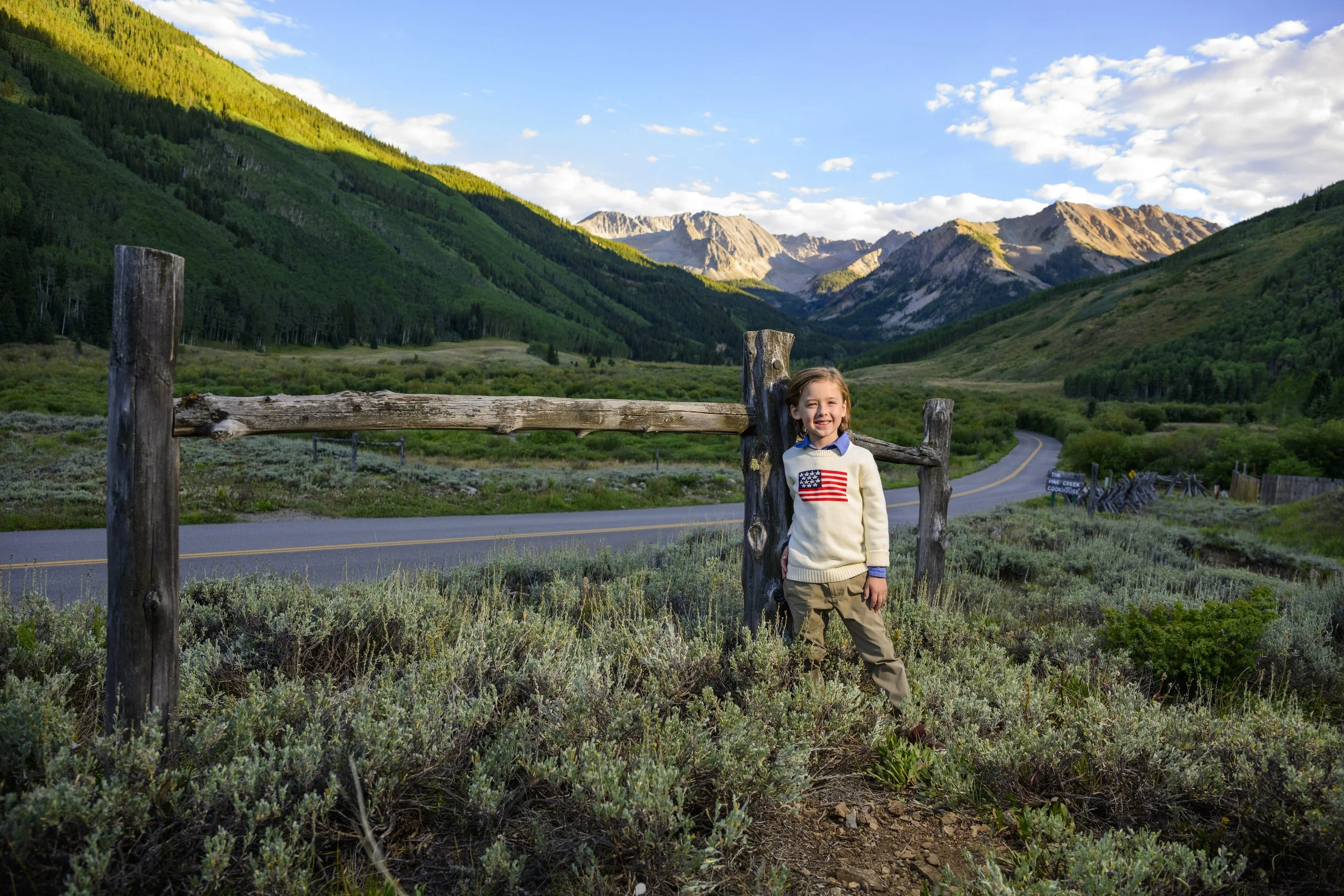 A young girl standing outdoors in front of a rustic wooden fence with a mountain valley and winding road in the background. She is smiling and wearing a sweater with an American flag patch, khaki pants, and brown shoes, surrounded by green landscape 