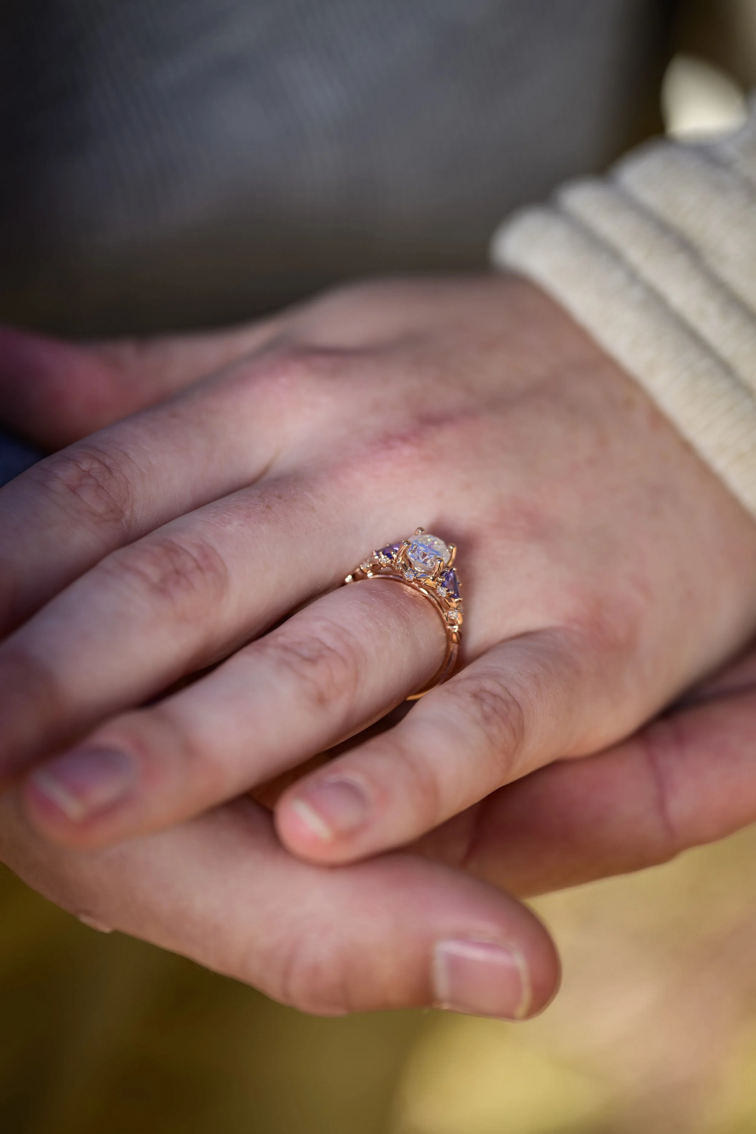 Close-up of a person's left hand with a gold ring featuring a large central gemstone and smaller stones, resting on the back of another person's hand.