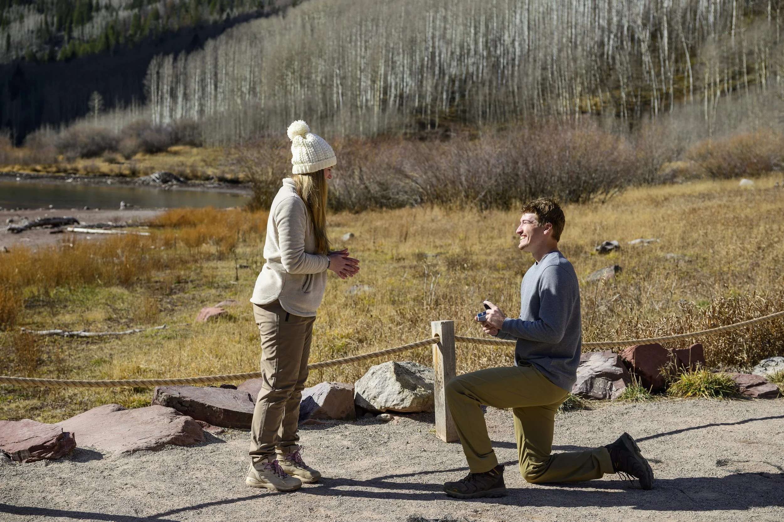 A man proposing to a woman outdoors in a scenic area with mountains and trees, while the woman looks surprised. The man is kneeling on one knee holding a ring box, and the woman is standing with her hands clasped.