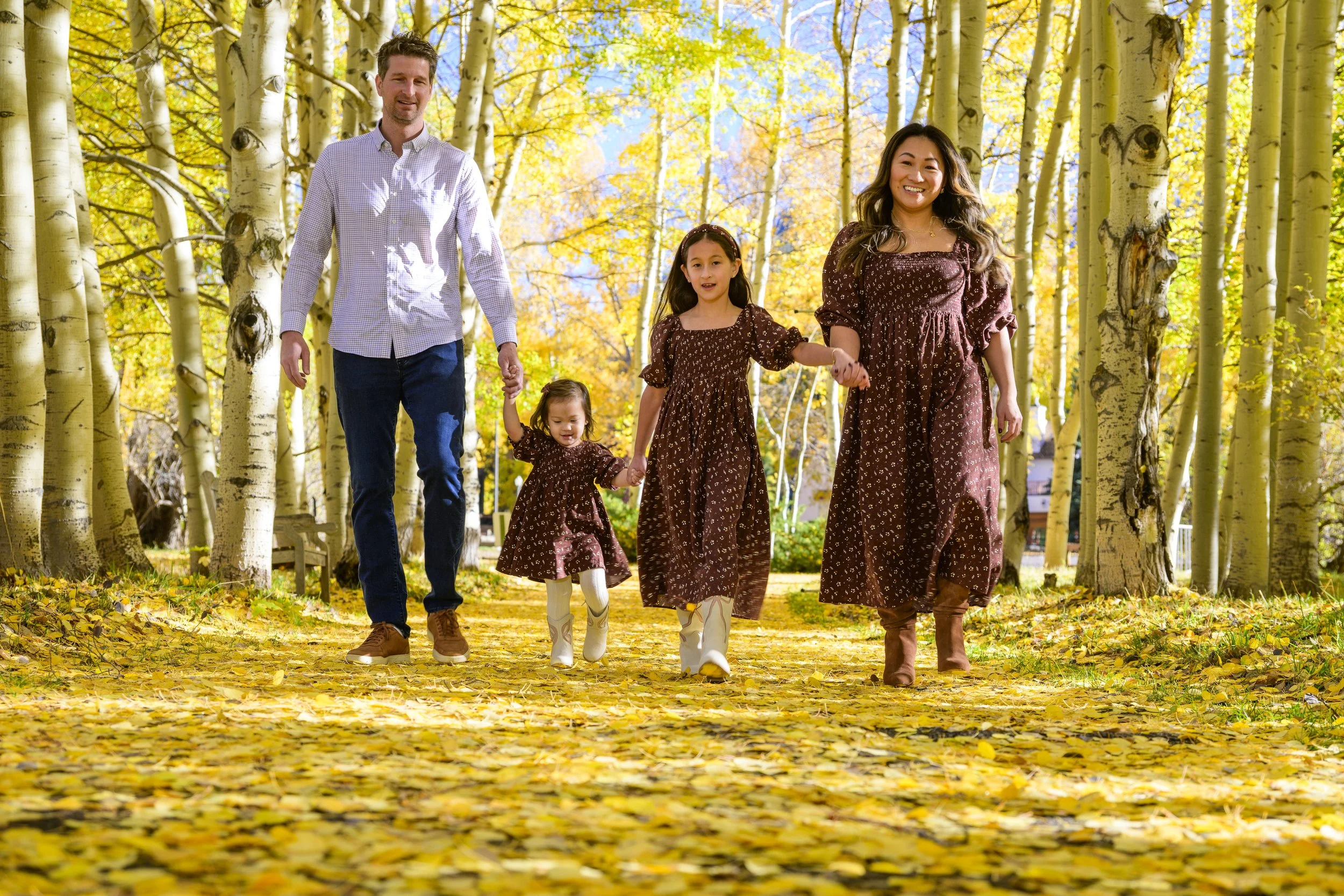 Family of four walking through a forest with yellow autumn leaves, holding hands, dressed in matching brown dresses and casual clothes.