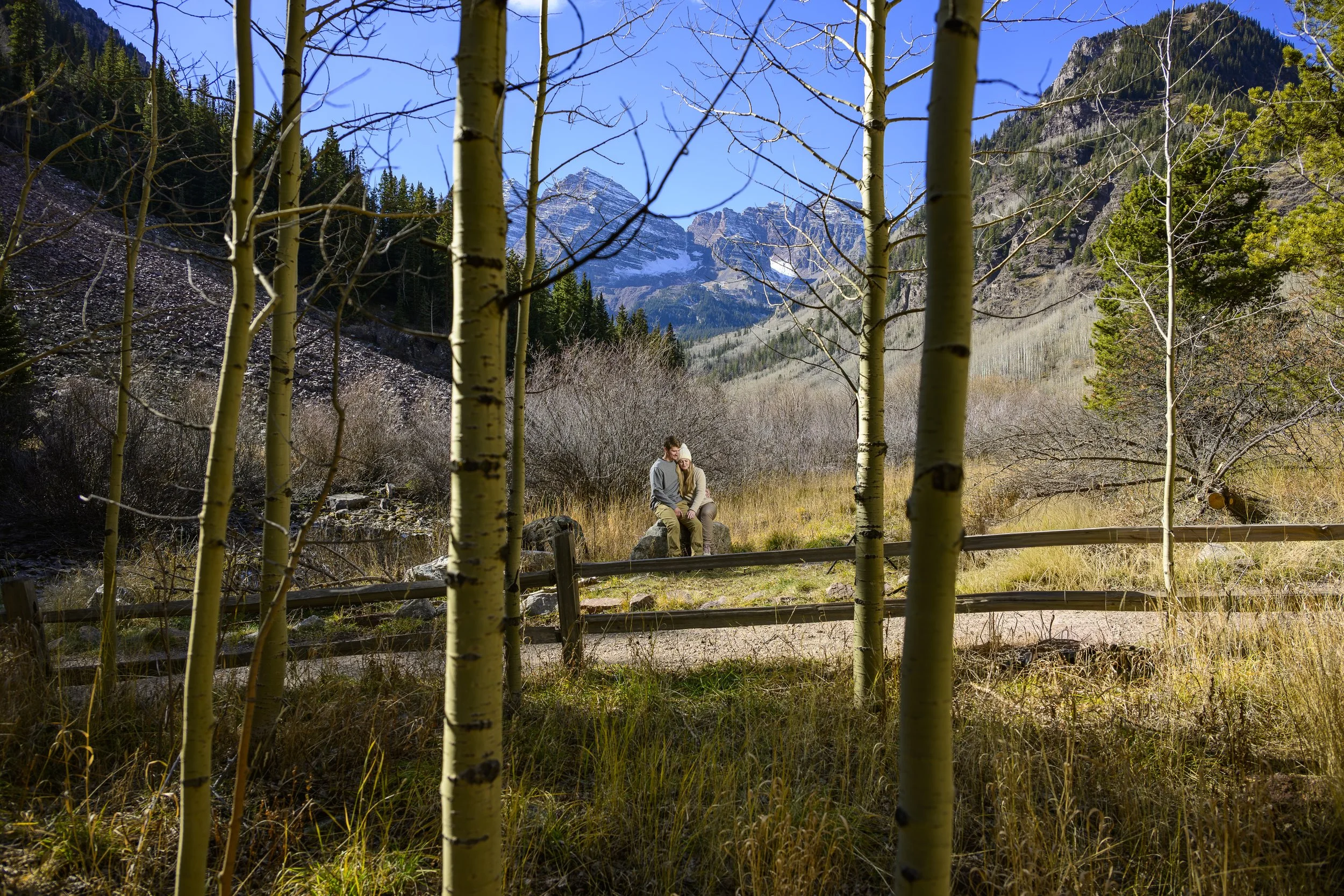 A couple sitting on a rock in a mountain valley, framed by yellow aspens and a wooden fence, with mountains and a clear blue sky in the background.