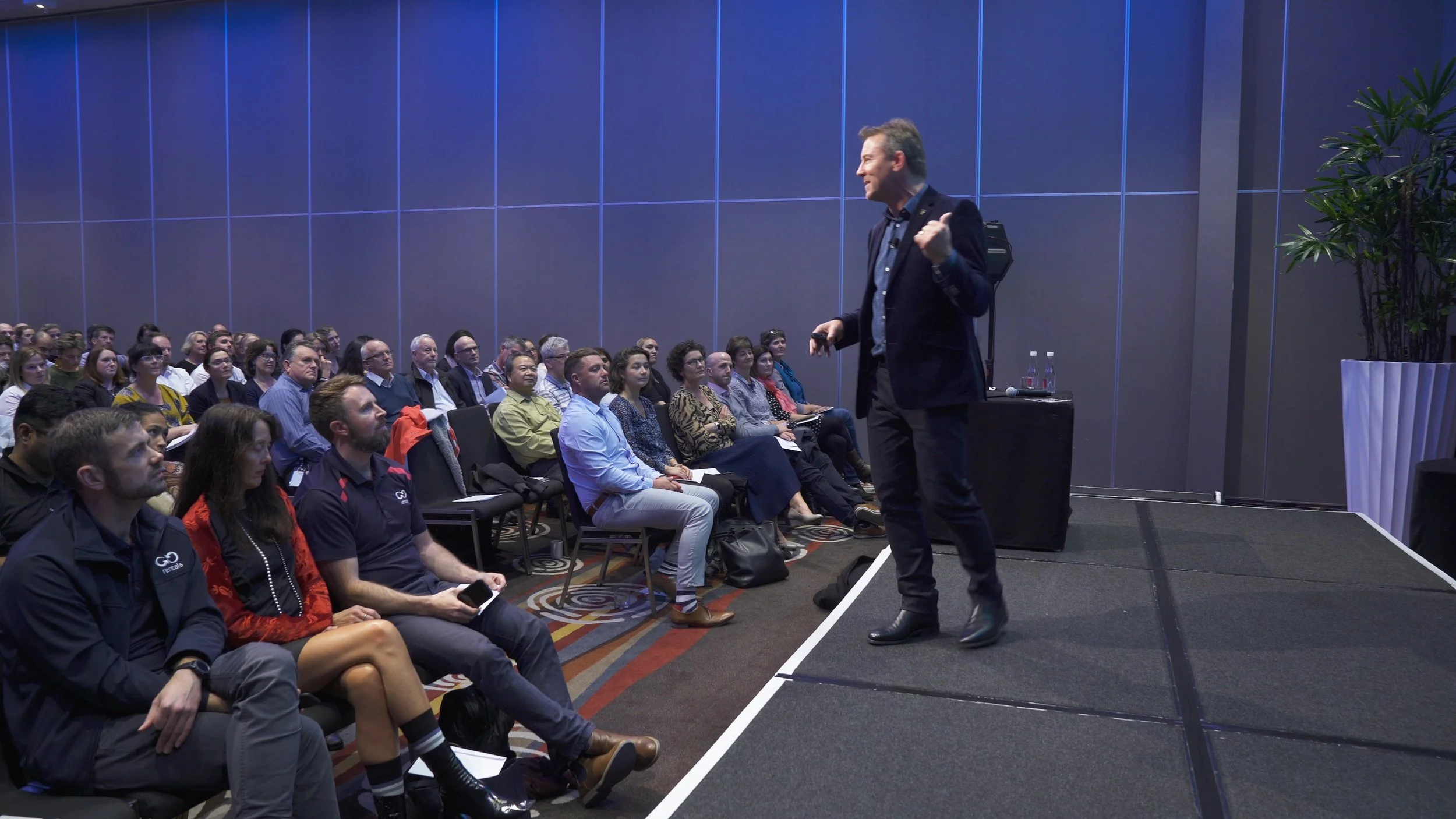 A man giving a presentation on a stage in front of a seated audience in a conference room.