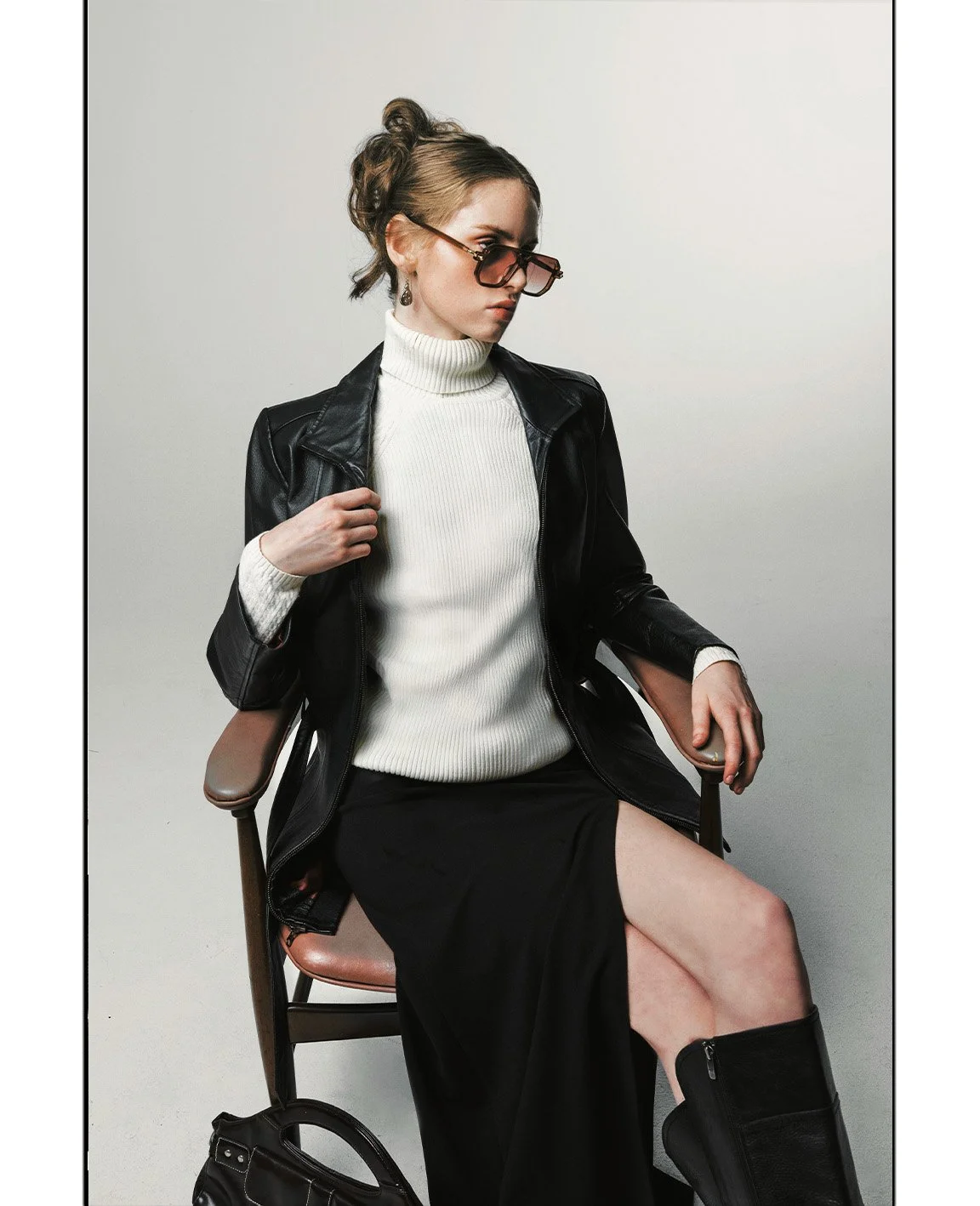 A woman with curly hair styled in an updo, wearing brown sunglasses, a white turtleneck sweater, a black leather jacket, a long black skirt, and black knee-high boots, sitting on a wooden chair with her legs crossed, holding the armrest. There is a b