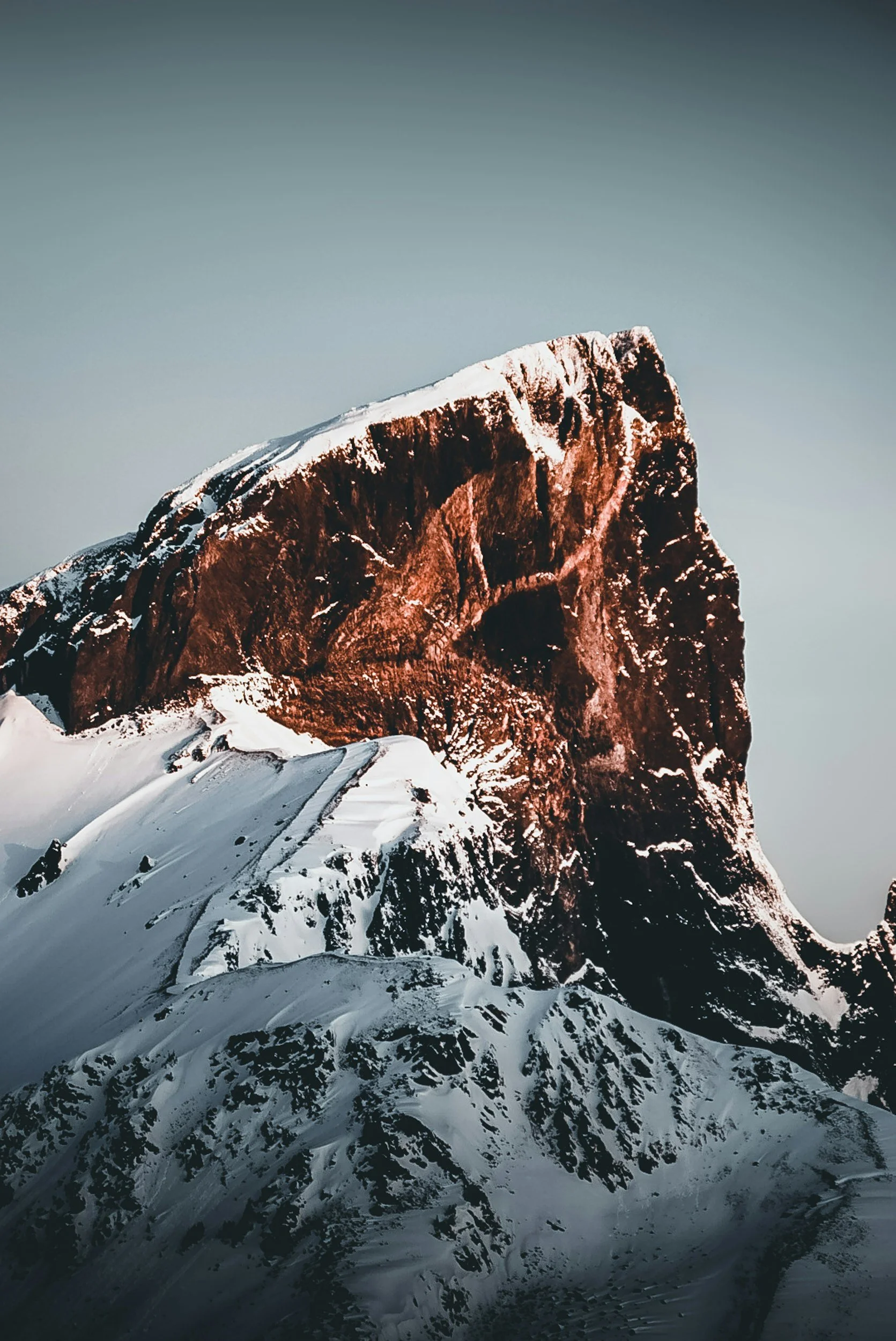 Snow-covered mountain peak with reddish-brown coloration on the exposed rock, under a clear pale blue sky.