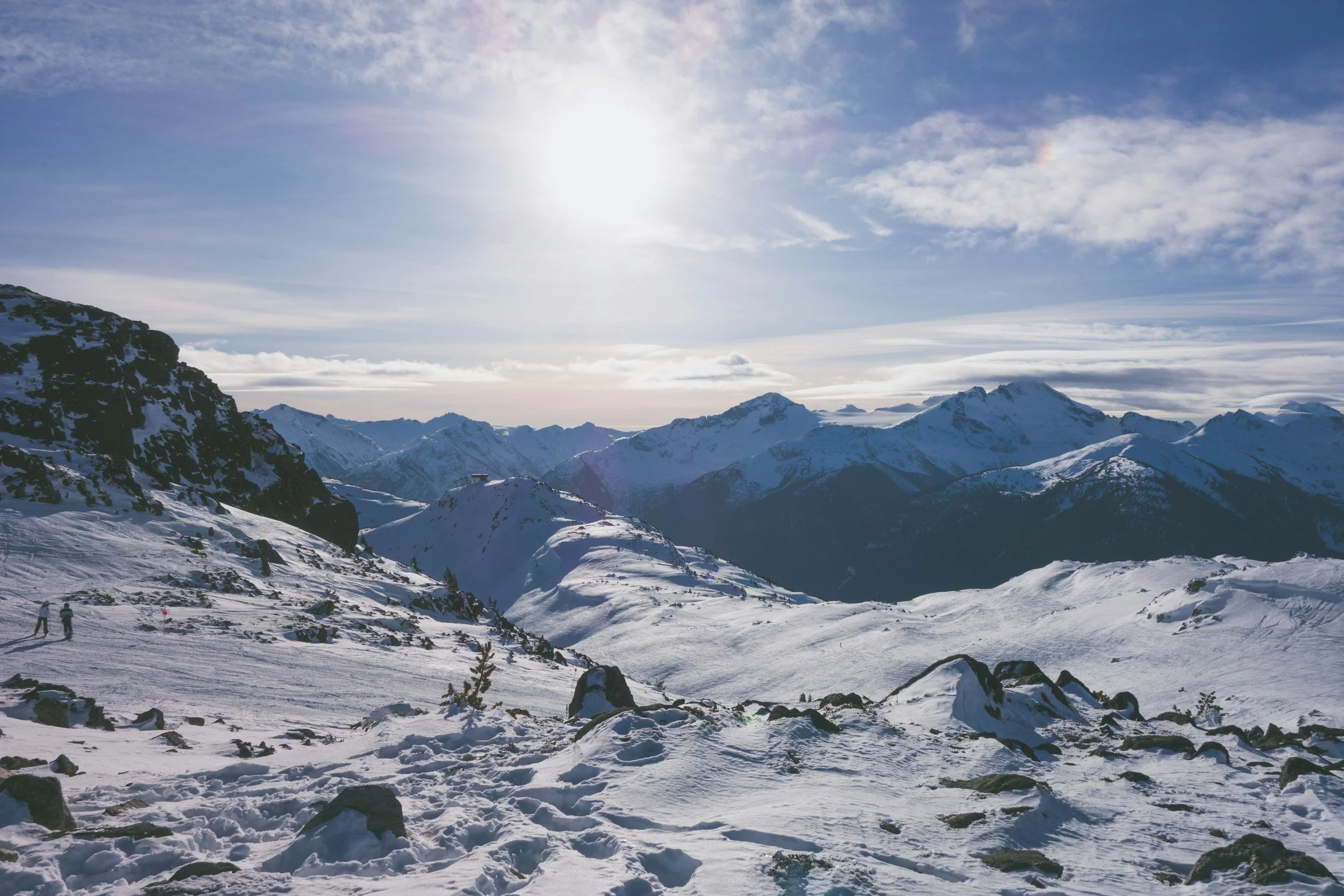Snow-covered mountain landscape with slopes, peaks, and a bright sun in a partly cloudy sky. Sparse trees and rocks are visible on the ground.