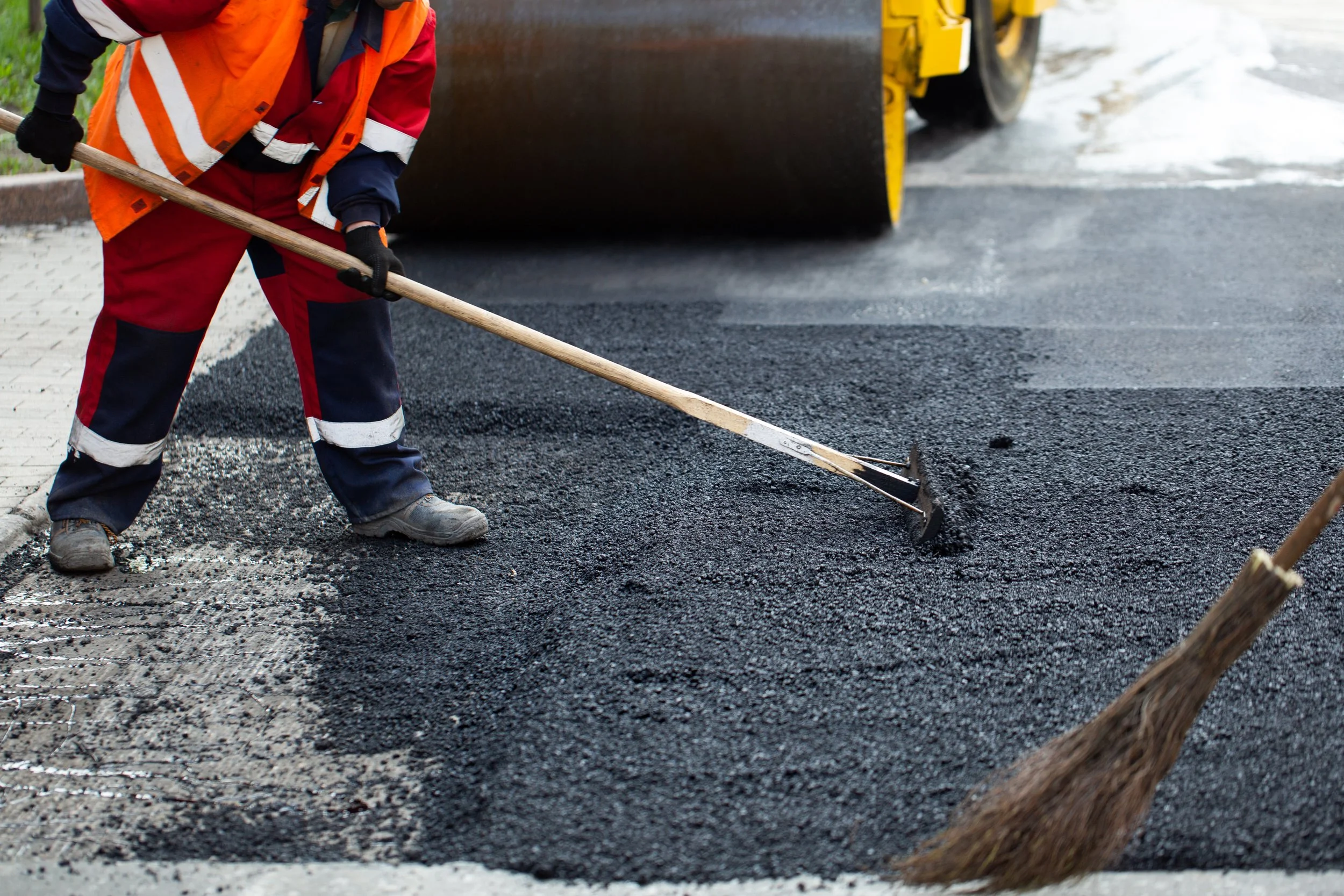Construction worker spreading asphalt with a rake on a road, with a broom and a steamroller in the background.