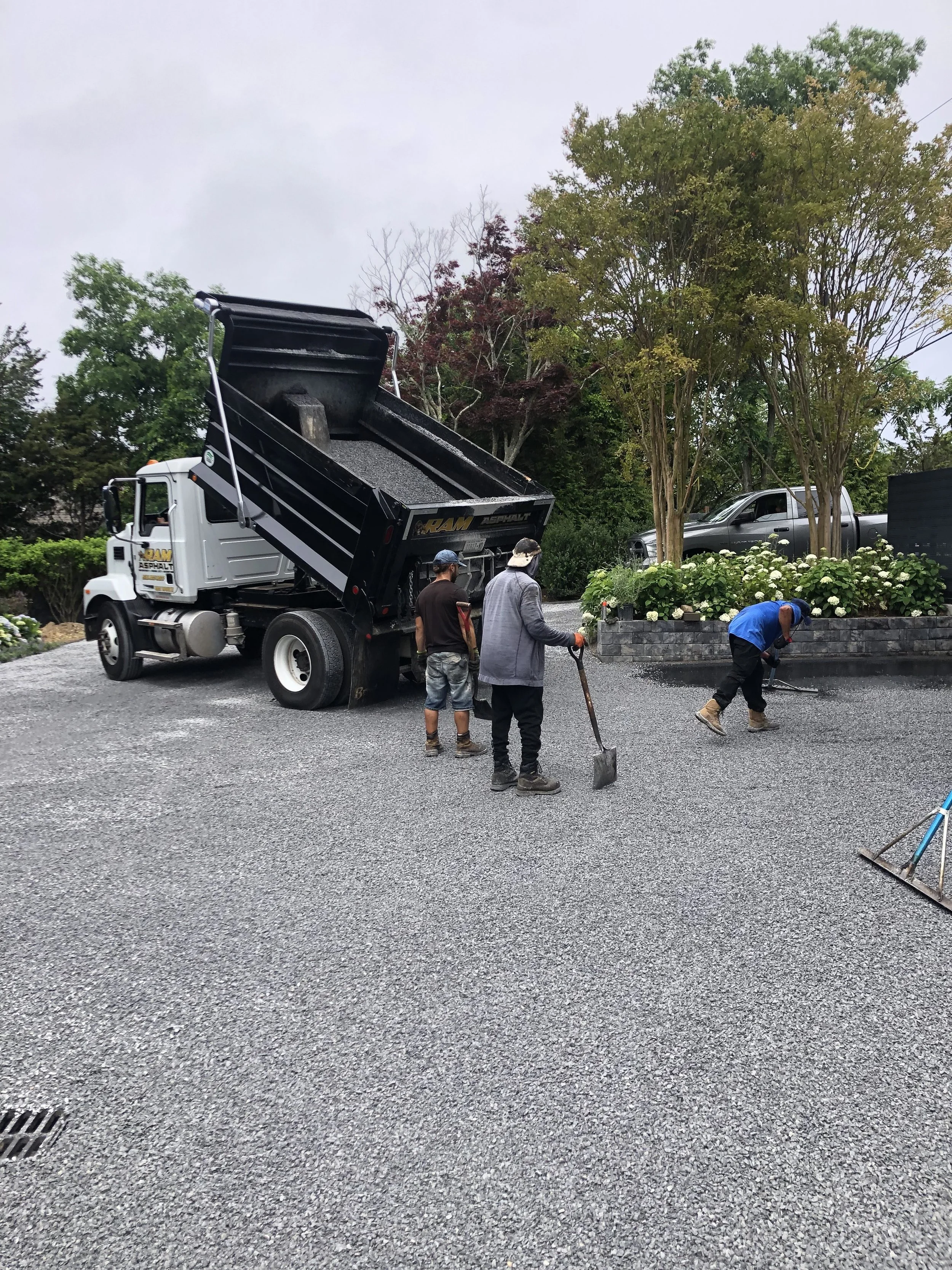 Workers laying gravel from a dump truck on a driveway, using shovels and rakes, with trees and a parked car in the background.