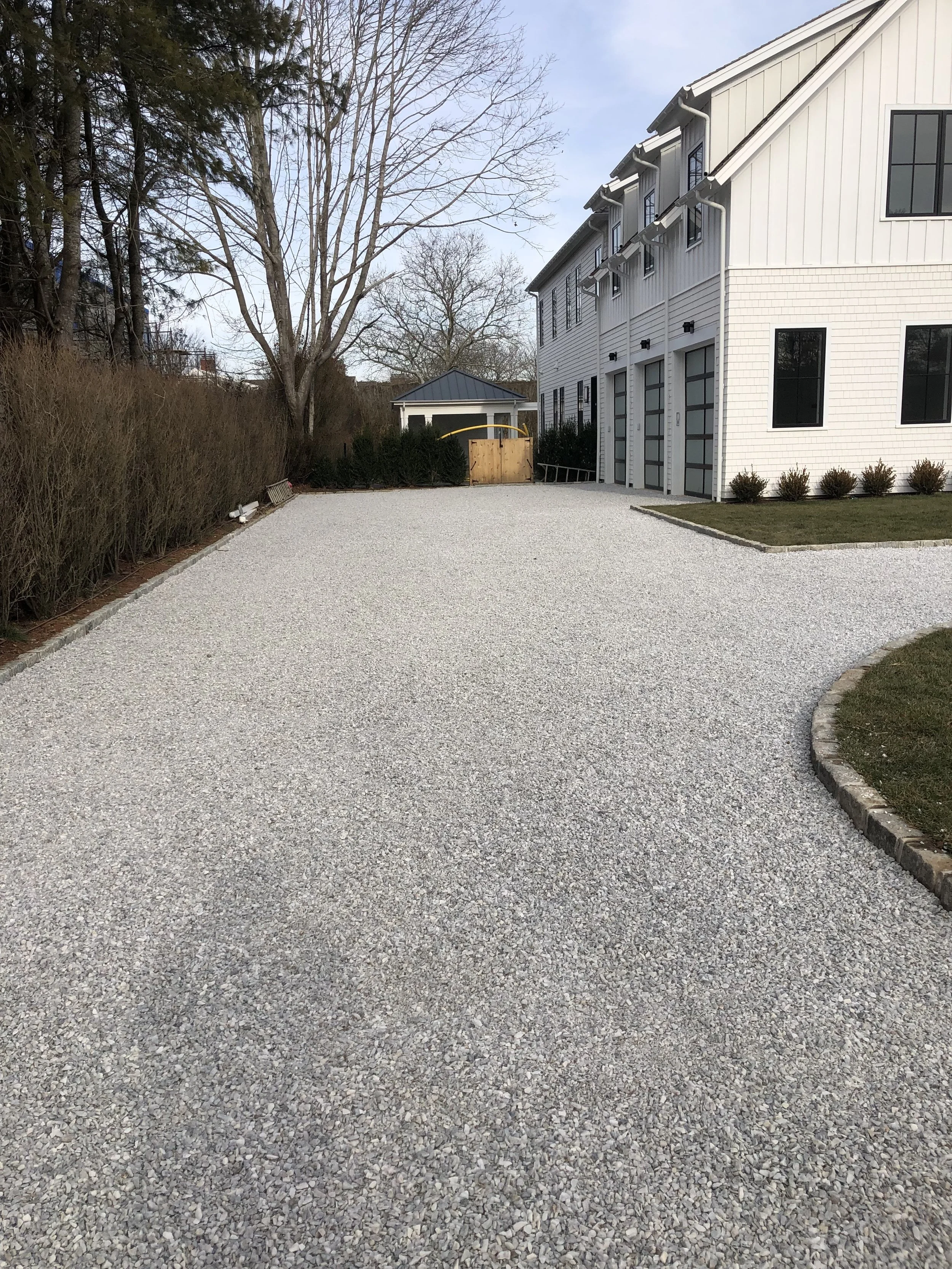 Gravel driveway in front of a modern white house with trees and shrubs around it.