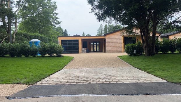 Modern house with a gravel driveway, surrounded by green grass and trees, featuring a blue portable toilet on the side.