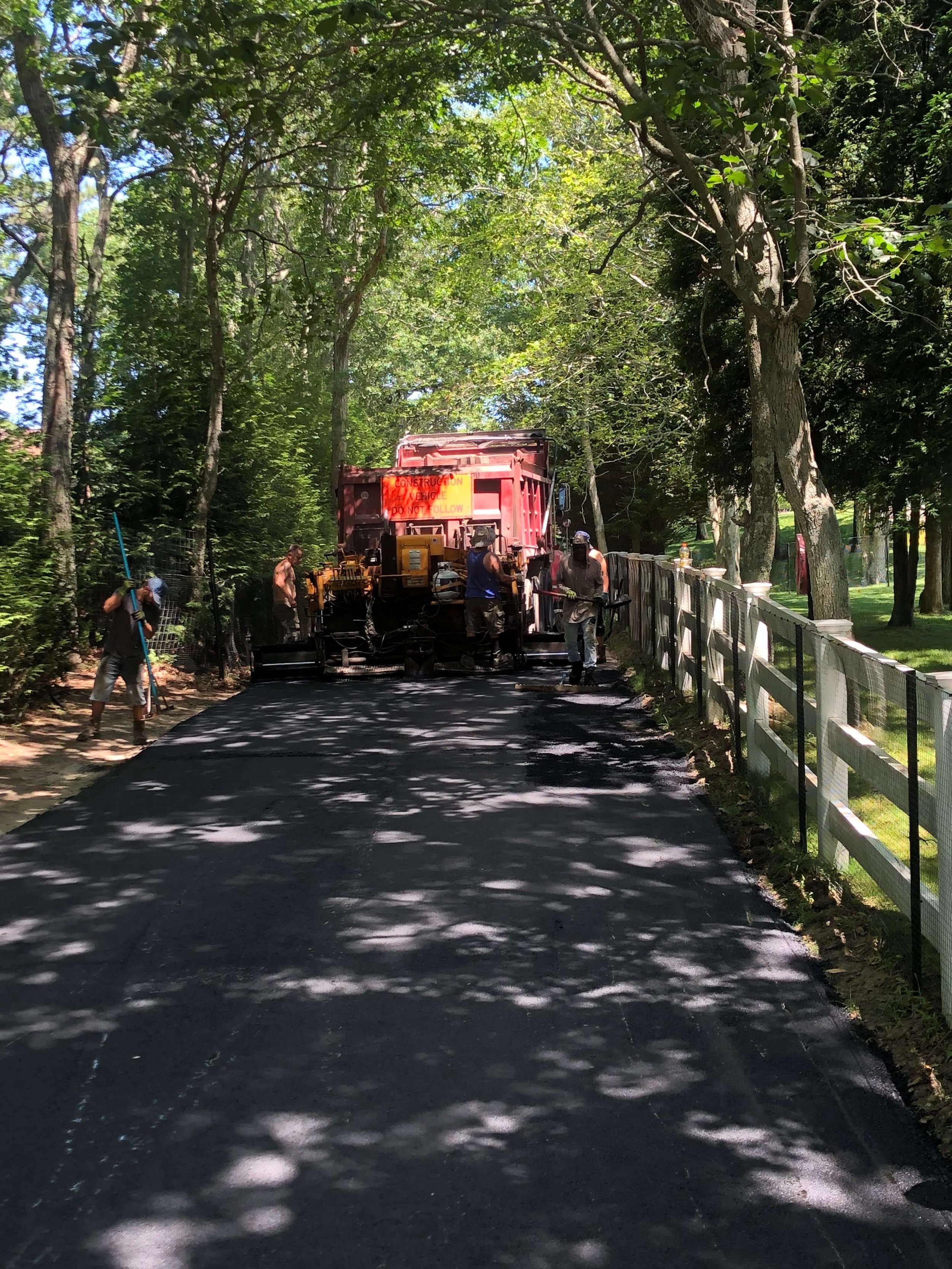 Workers paving a road in a wooded area with trees and a white fence on the right side.