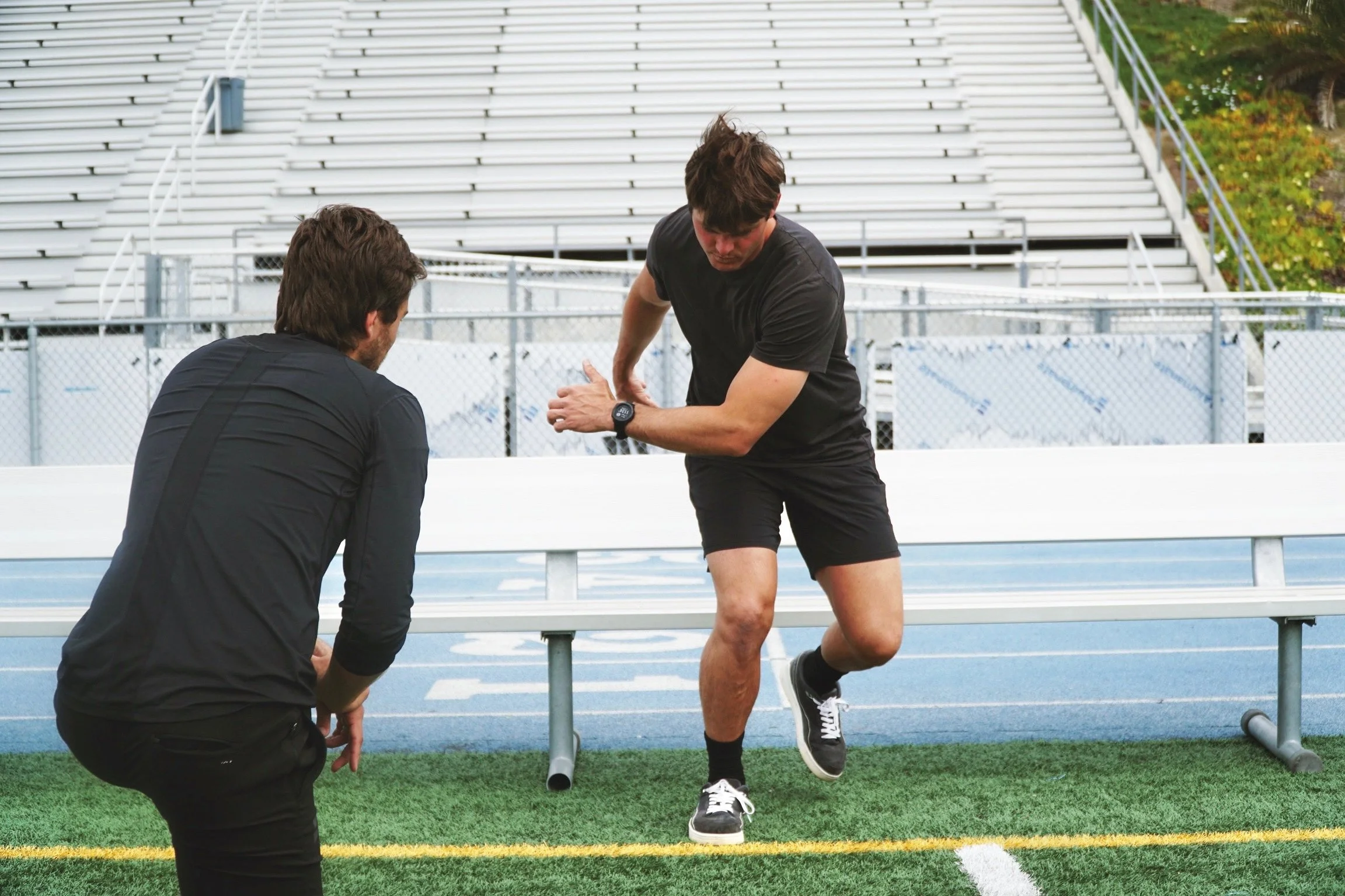 Two men training on a football field; one running, the other observing, with bleachers in the background.