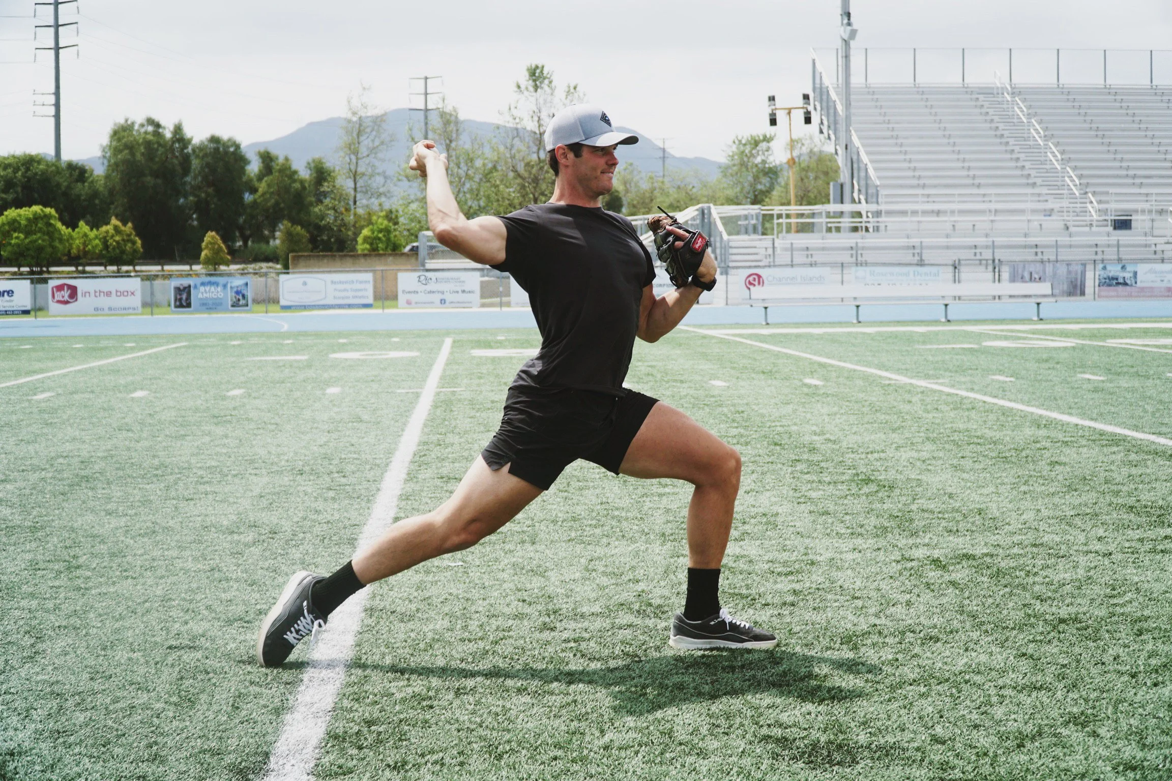 Man practicing baseball pitching on outdoor field, wearing black shorts and shirt, holding a baseball mitt, with empty bleachers in the background.